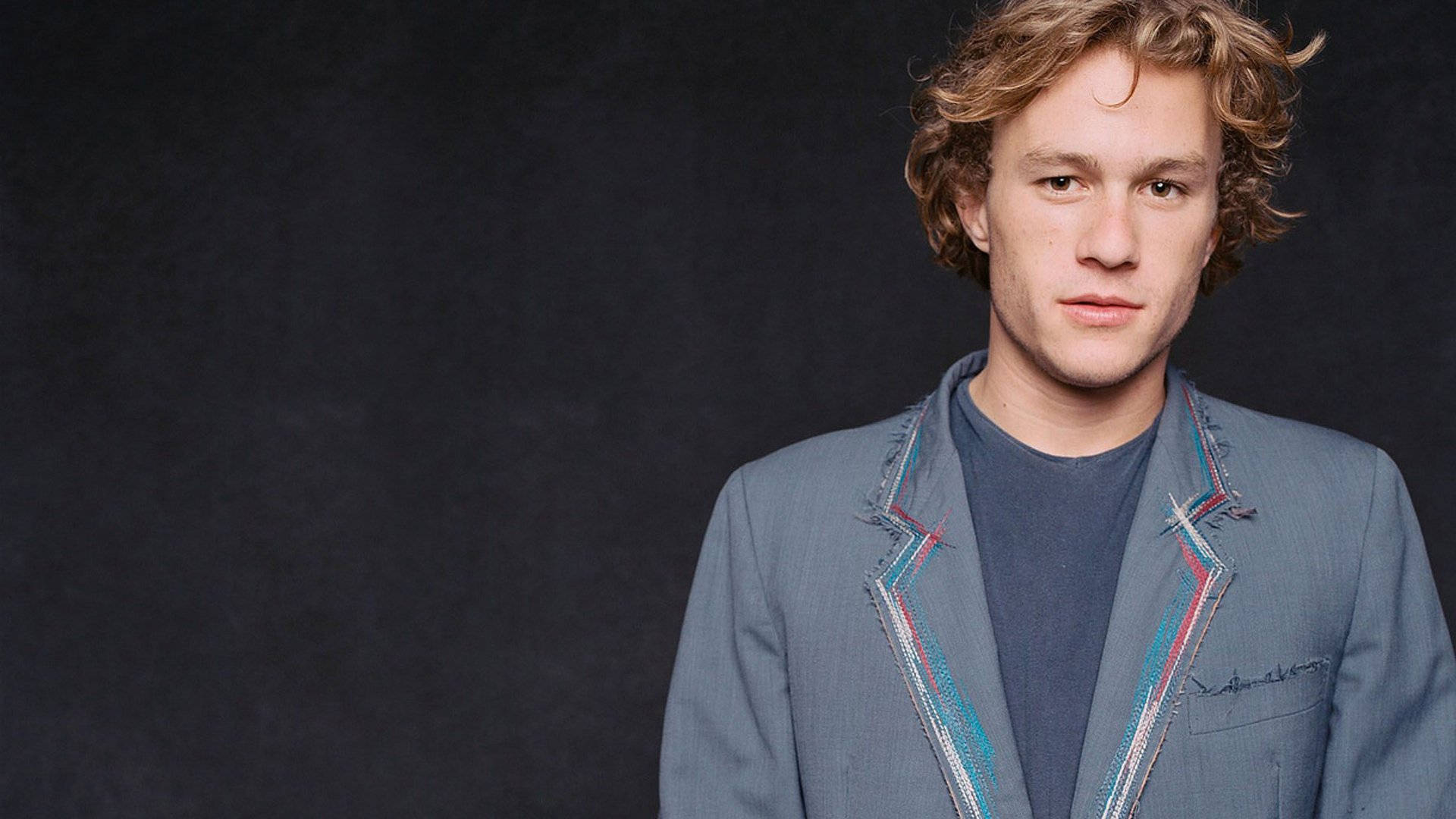 Heath Ledger 桌布 壁紙 — 一位年輕男子，一頭凌亂的金髮，身穿灰色西裝外套，帶有彩色條紋飾邊，背景為深色紋理。