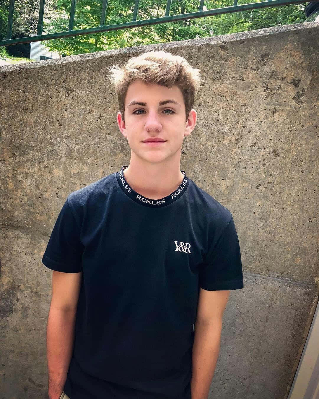 teenager 桌布 壁紙 — 一位年輕男子，一頭凌亂金髮，在明亮的日光下靠著風化的混凝土牆站立。