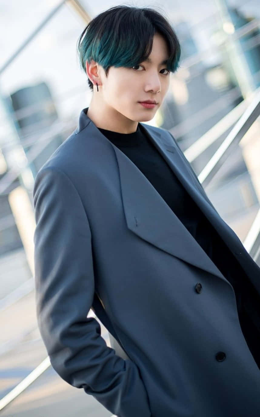 Jungkook 桌布 壁紙 — 一位年輕男子，帶有藍綠色髮尾和灰色外套，自信地站在模糊的現代玻璃建築背景前。