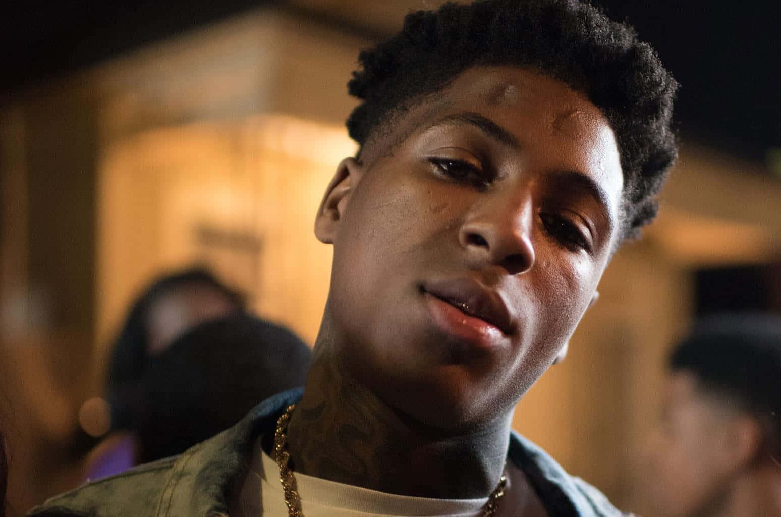 Youngboy 桌布 壁紙 — 一位年輕男子，捲髮，頸部有刺青，在溫暖的室內環境中若有所思地凝視著。