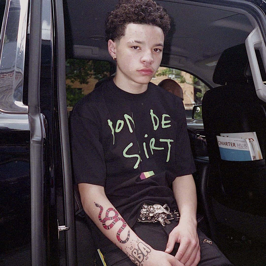 Lil Mosey 桌布 壁紙 — 一位年輕男子，捲髮且有刺青，坐在黑色SUV的後座，身穿帶有綠色文字的黑色T恤。