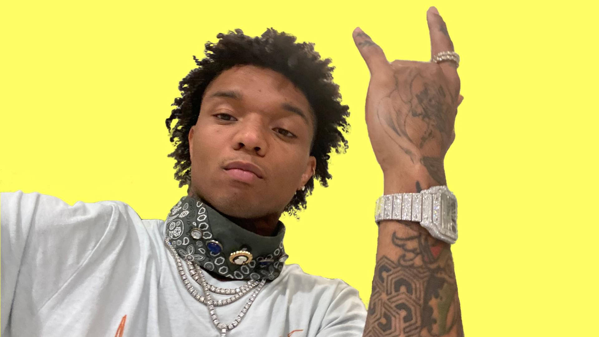 Swae Lee 桌布 壁紙 — 一位年輕男子，捲髮和刺青，在明亮的黃色背景前自信地擺姿勢，做出搖滾手勢，戴著頭巾和鑲鑽手錶。