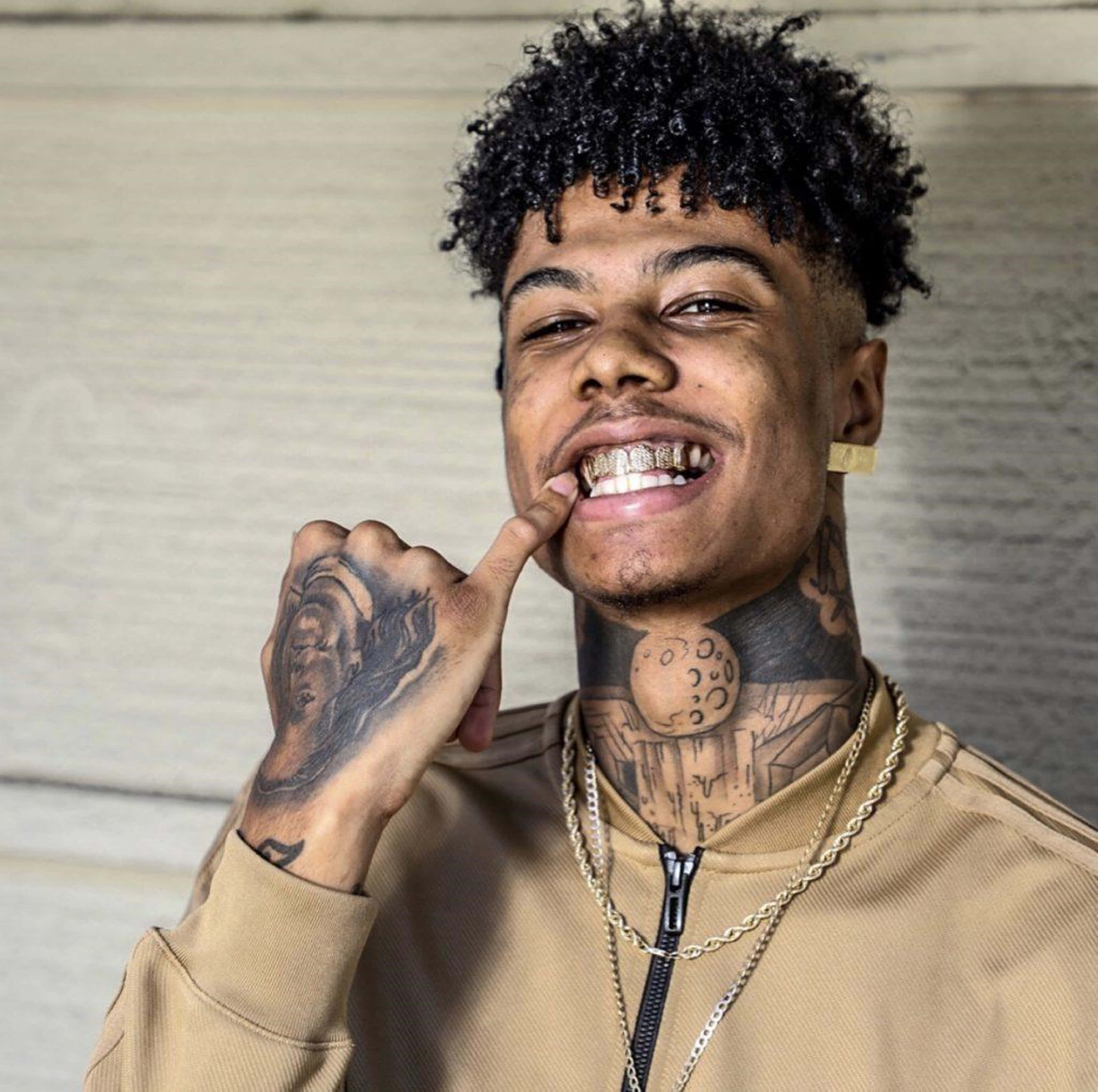 4K Rapper 桌布 壁紙 — 一位年輕男子，捲髮和刺青，微笑著指著他的金色牙套，背景是木牆。