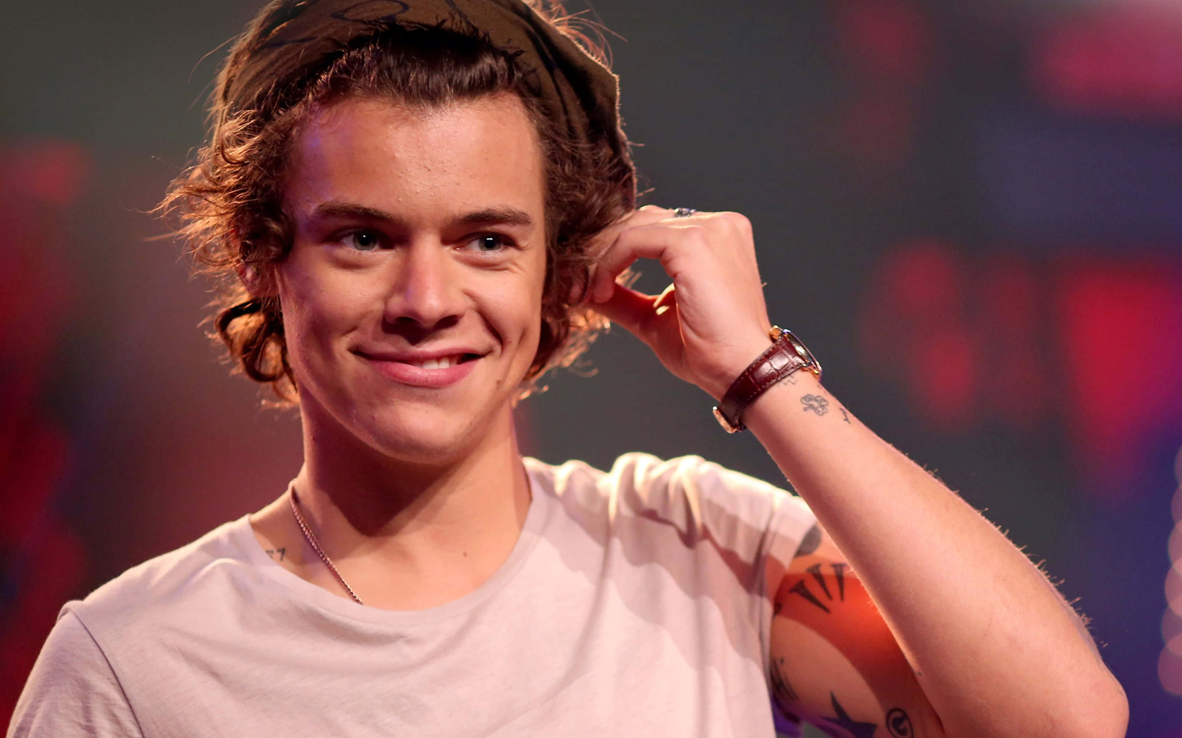 Harry Styles Laptop 桌布 壁紙 — 一位年輕男子，捲髮戴頭帶，在色彩繽紛的舞台上微笑觸摸耳朵。