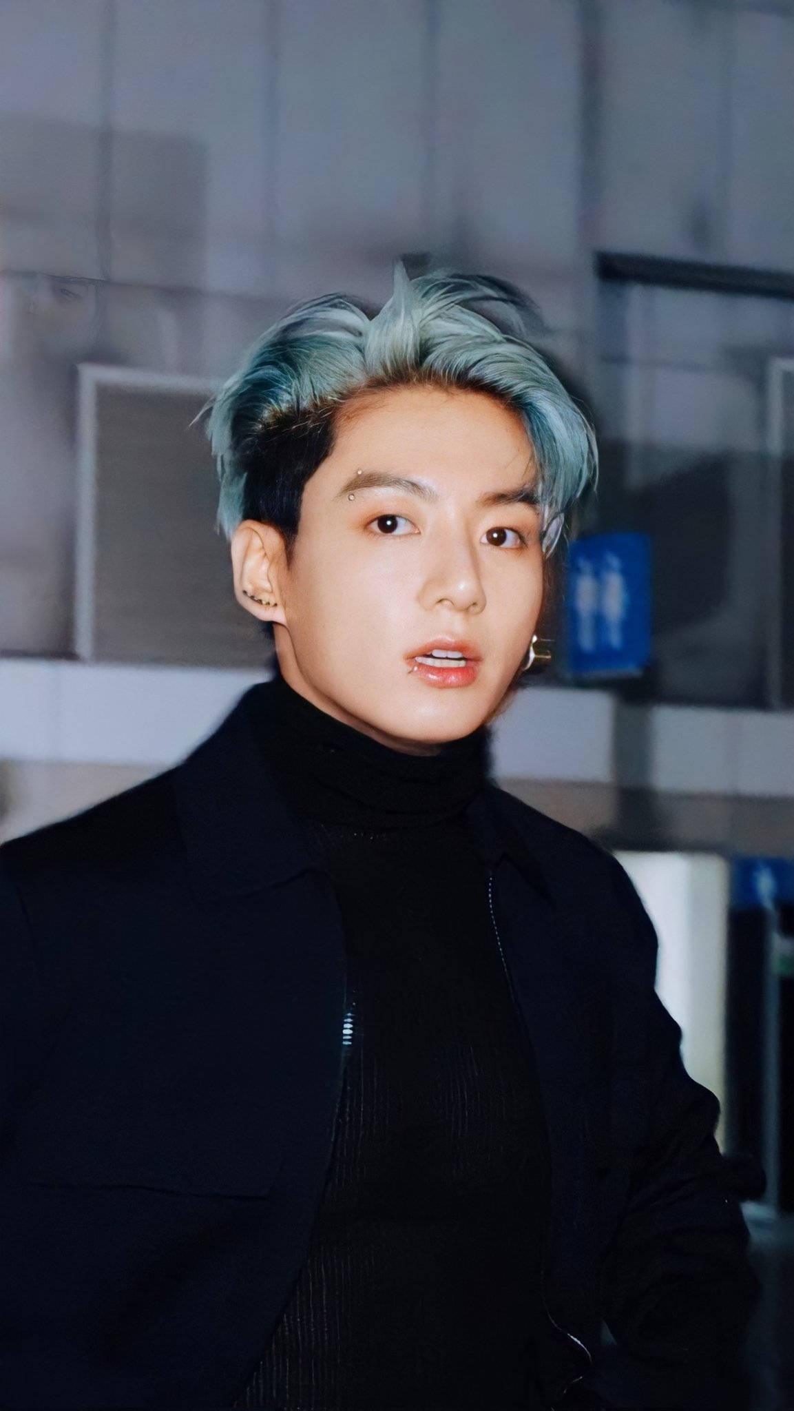 BTS 桌布 壁紙 — 一位年輕男子，擁有引人注目的藍色髮尖和深色服裝，在現代室內環境中直視鏡頭。