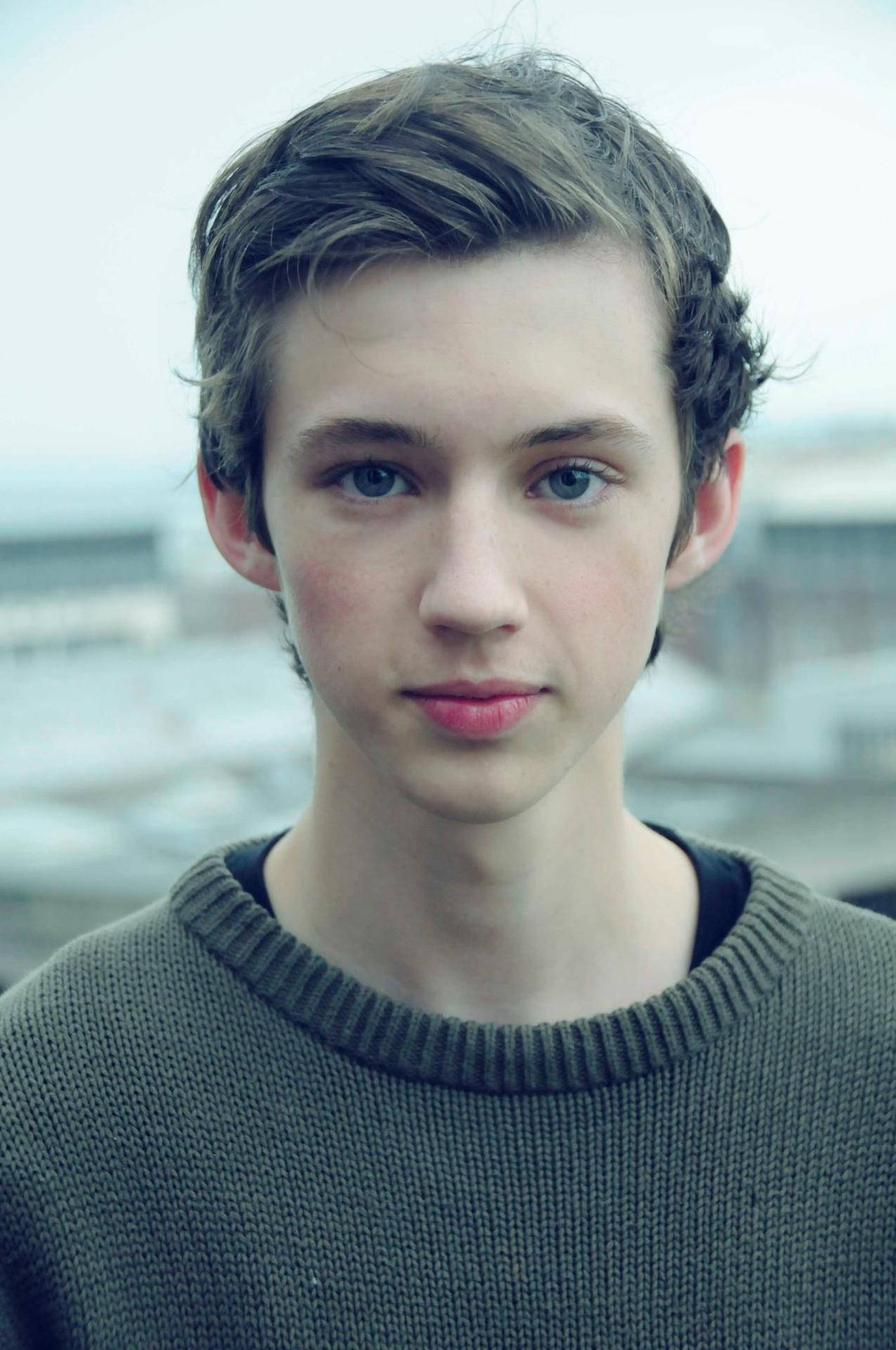 Sivan 桌布 壁紙 — 一位年輕男子，擁有引人注目的藍色眼睛和凌亂的棕色頭髮，穿著質感灰色毛衣，平靜地凝視著相機。