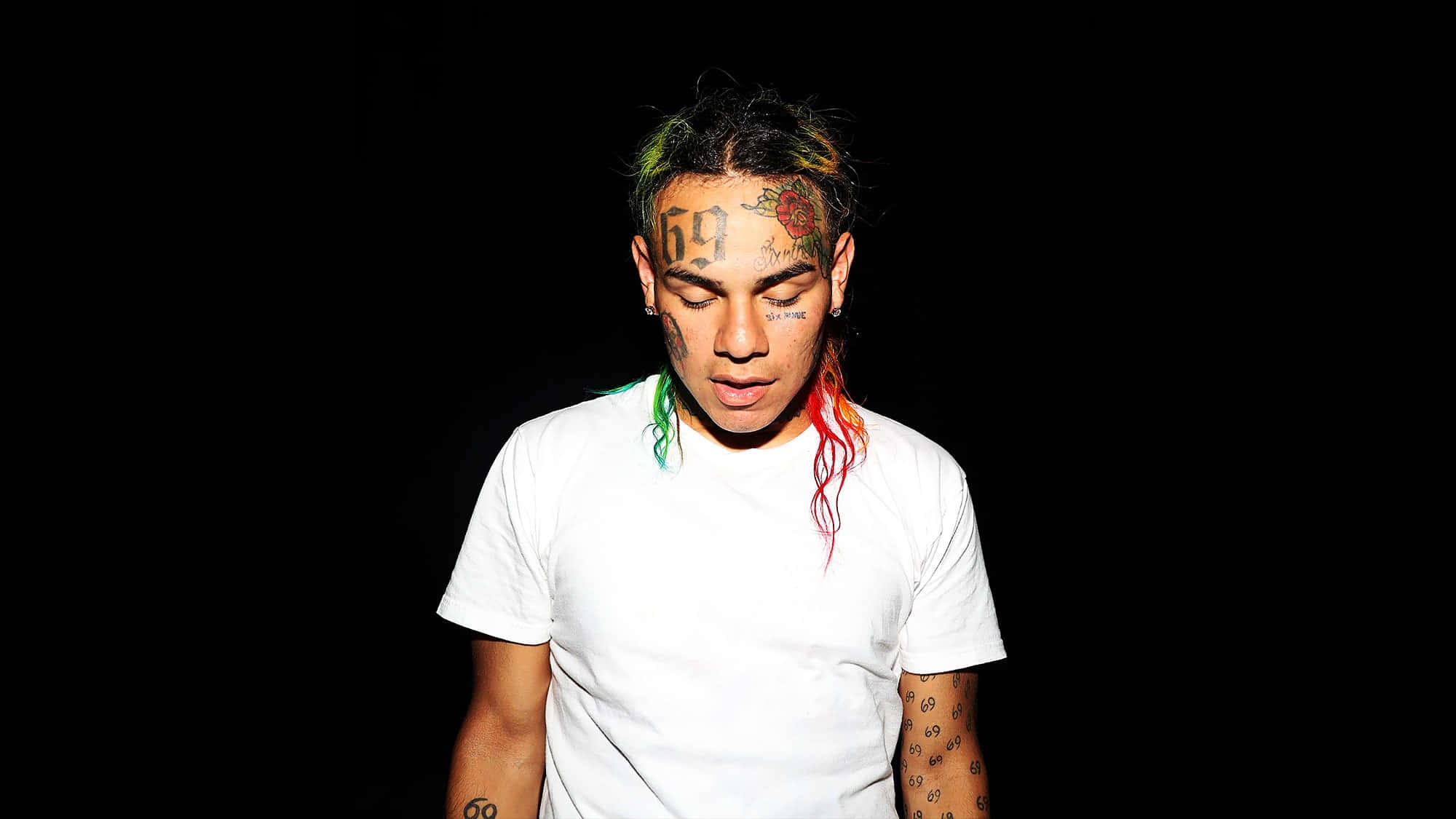 69 Rapper 桌布 壁紙 — 一位年輕男子，擁有色彩繽紛的頭髮和刺青，穿著白色T恤，在黑色背景前向下凝視。