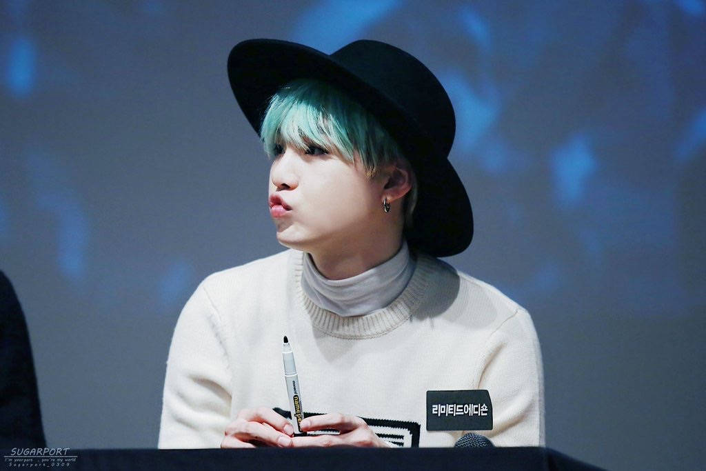 Bts Suga Cute 桌布 壁紙 — 一位年輕男子，擁有藍綠色頭髮和黑色帽子，坐在桌子旁，手持記號筆，背景是藍色模糊的。
