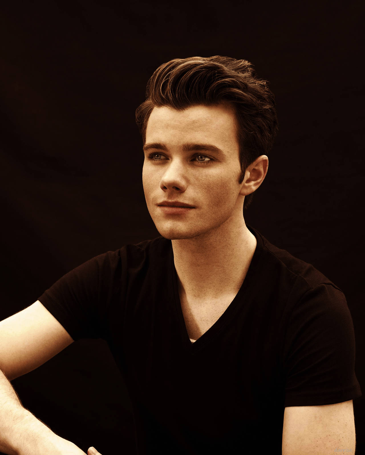 Chris Colfer 桌布 壁紙 — 一位年輕男子，擁有造型棕色頭髮和綠色眼睛，在深色背景下穿著黑色V領T恤，若有所思地凝視著。