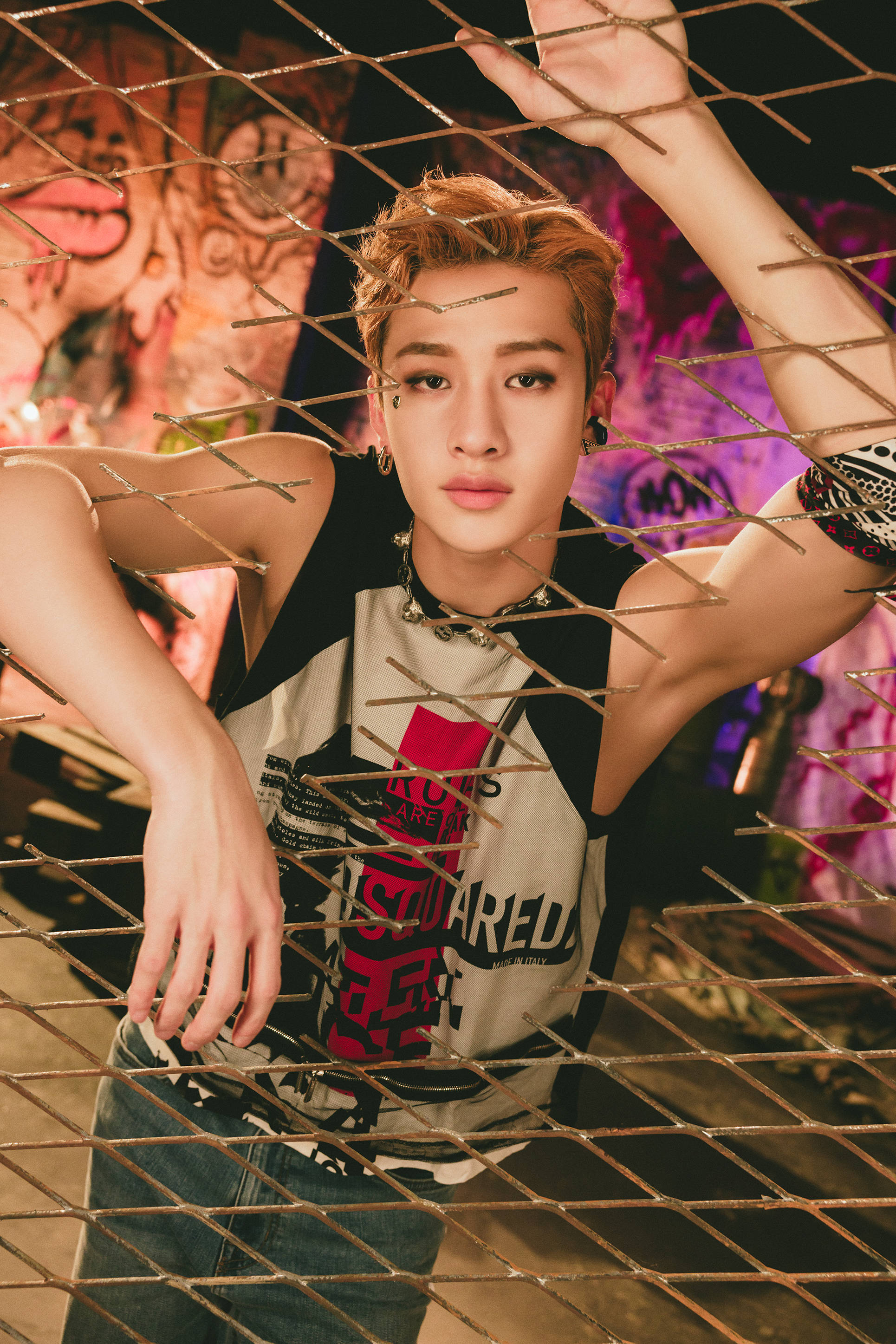 Stray Kids 桌布 壁紙 — 一位年輕男子，擁有造型頭髮和大膽配飾，在充滿活力的霓虹燈光下，在塗鴉牆前的鐵絲網後面擺姿勢。