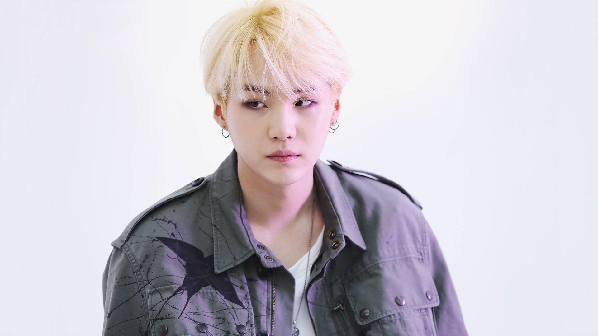 Suga Bts 桌布 壁紙 — 一位年輕男子，擁有鉑金色頭髮和深色星星圖案外套，在純白背景前凝視著。