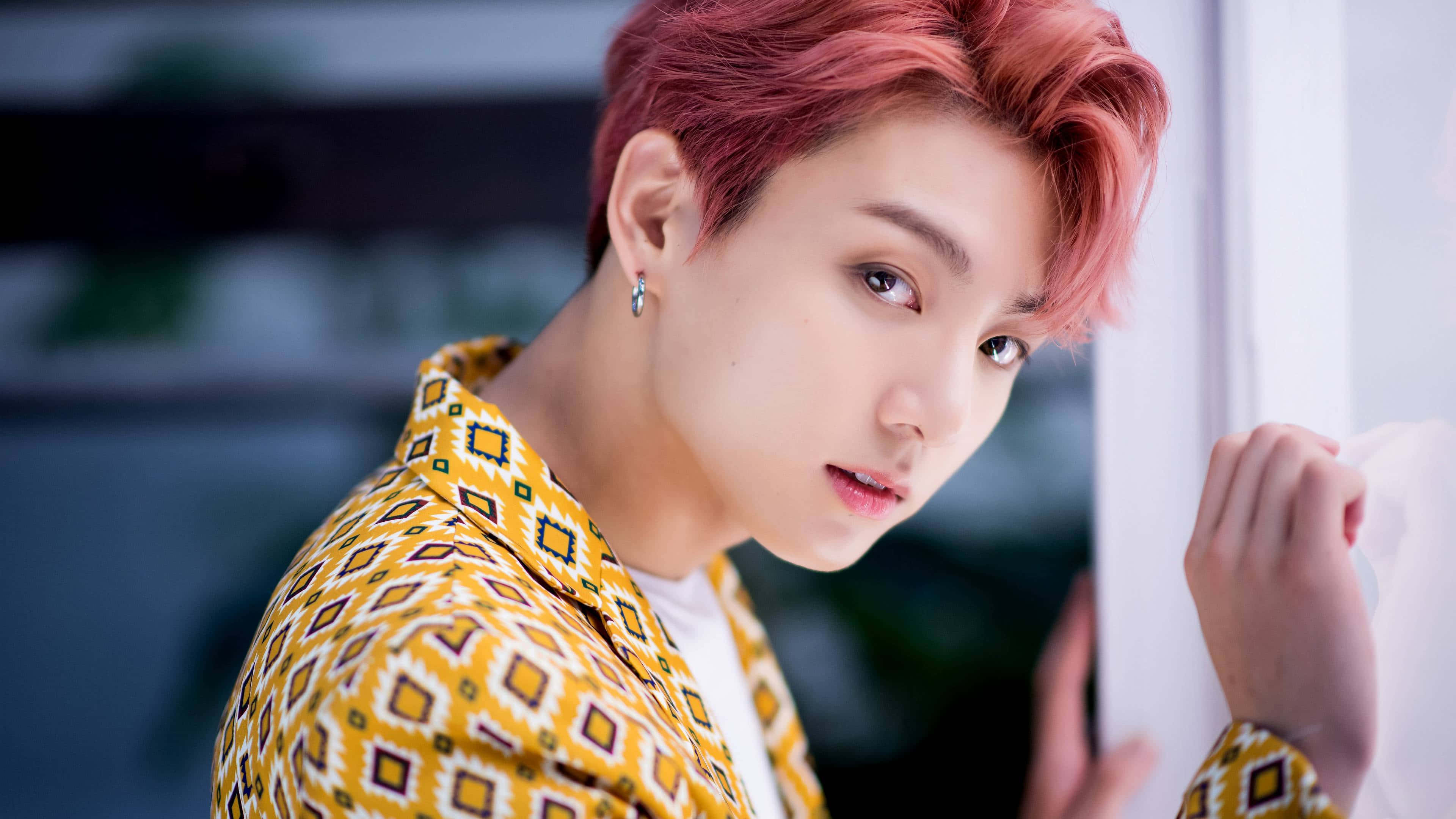 Jungkook 桌布 壁紙 — 一位年輕男子，擁有鮮豔的粉紅色頭髮和幾何圖案的黃色襯衫，靠著白牆專注地凝視。