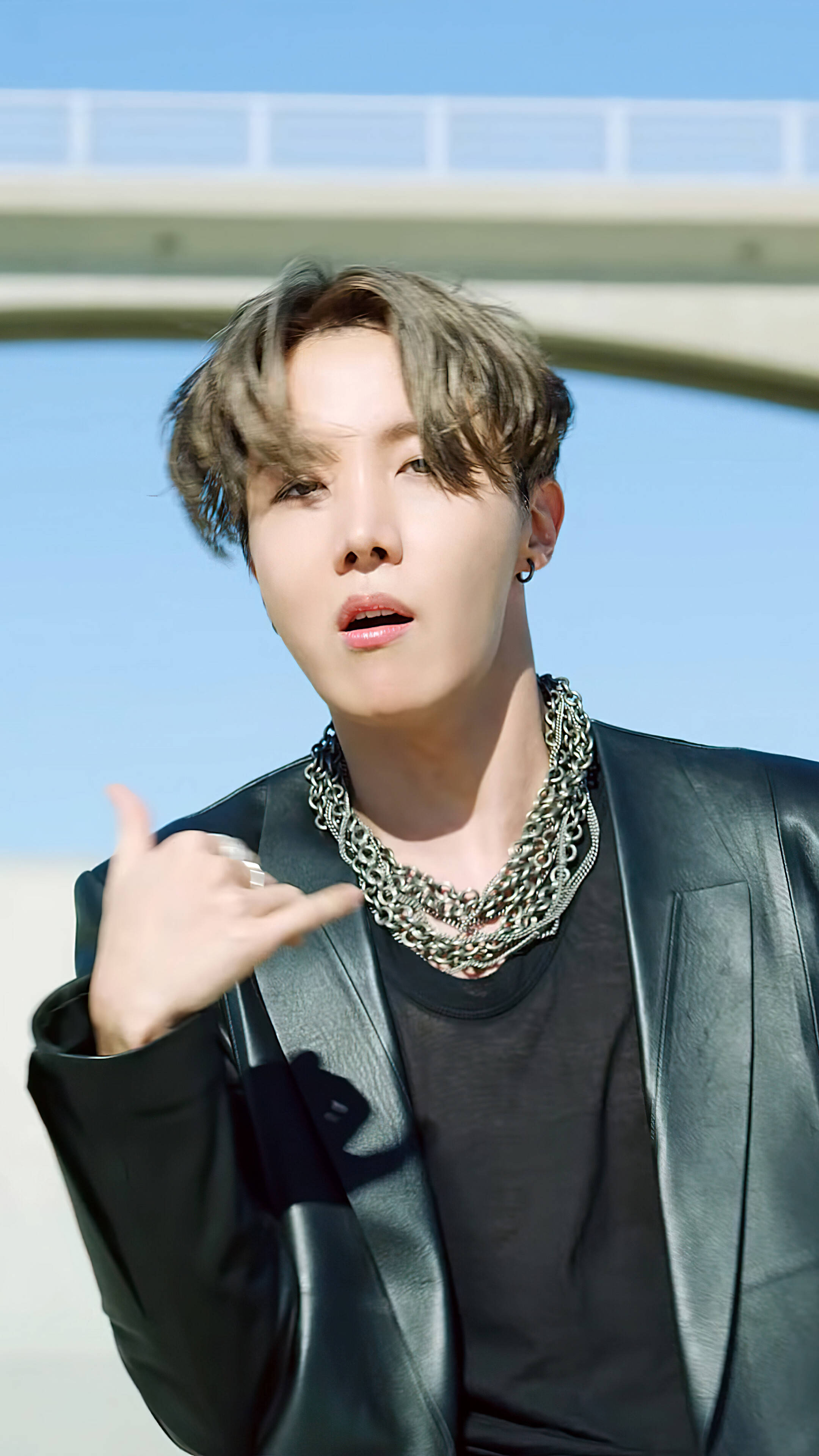 BTS 桌布 壁紙 — 一位年輕男子，梳著時尚髮型，佩戴多層銀鍊，在橋下以清澈藍天為背景自信地擺姿勢。