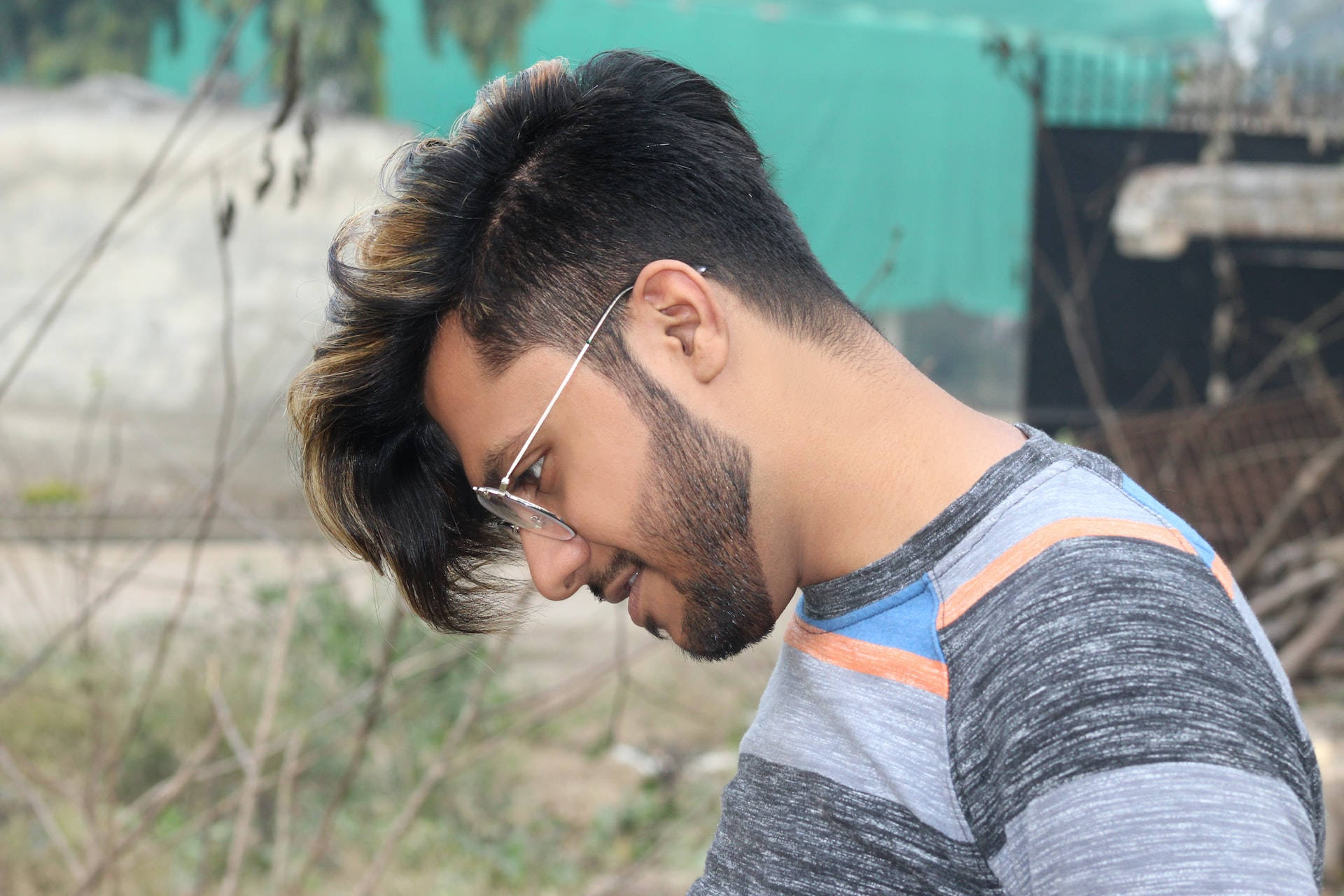 Haircut 桌布 壁紙 — 一位年輕男子，梳著時尚髮型並戴著眼鏡，在自然戶外環境中低頭注視。