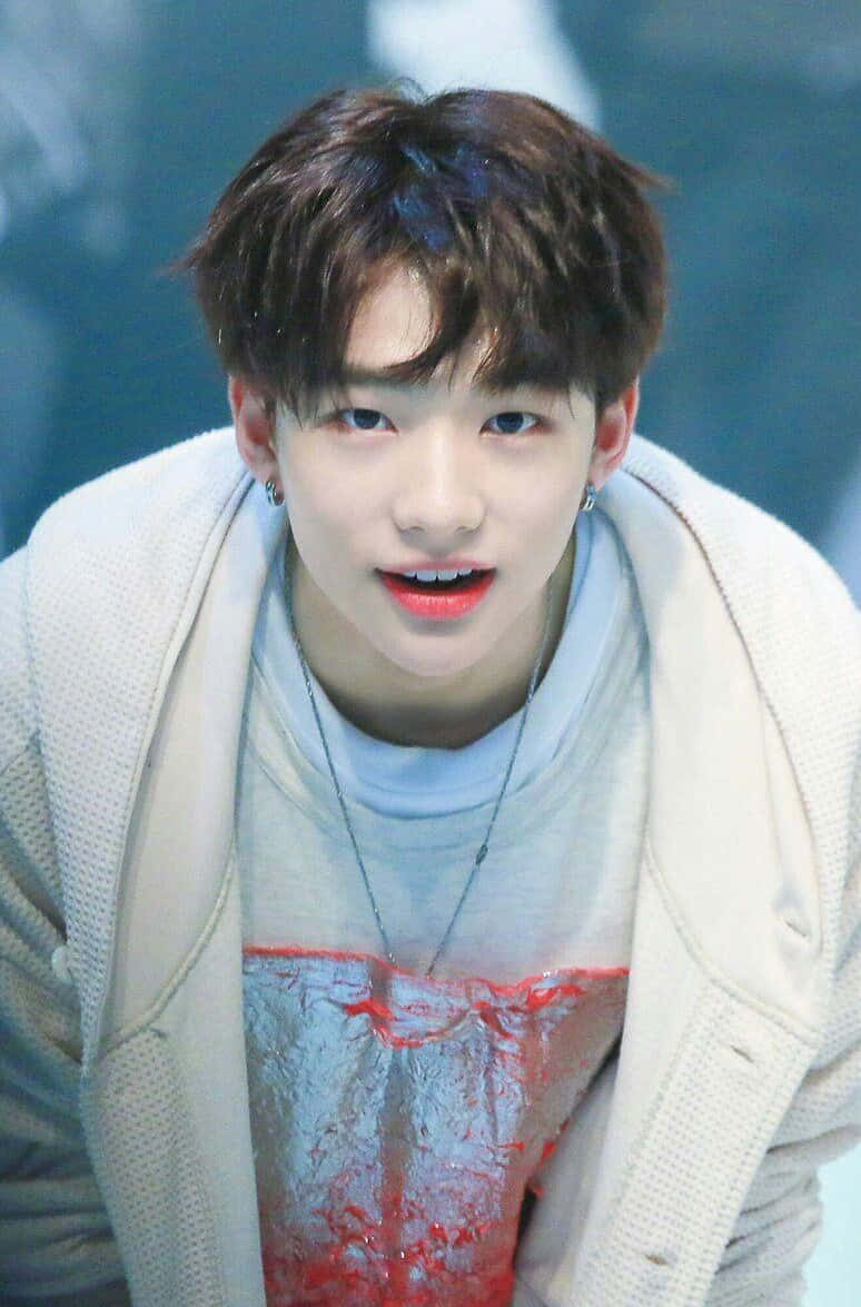 hyunjin 桌布 壁紙 — 一位年輕男子，棕色亂髮，塗著鮮紅色唇膏，穿著米色連帽上衣內搭圖案T恤，頑皮地微笑。