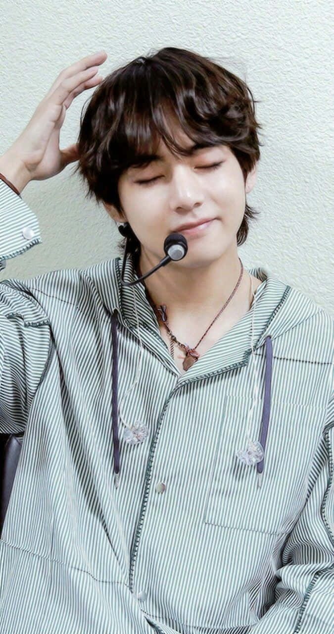 BTS V 動圖 桌布 壁紙 — 一位年輕男子，棕色亂髮，微笑著輕觸頭部，身穿條紋連帽衫和耳機麥克風。