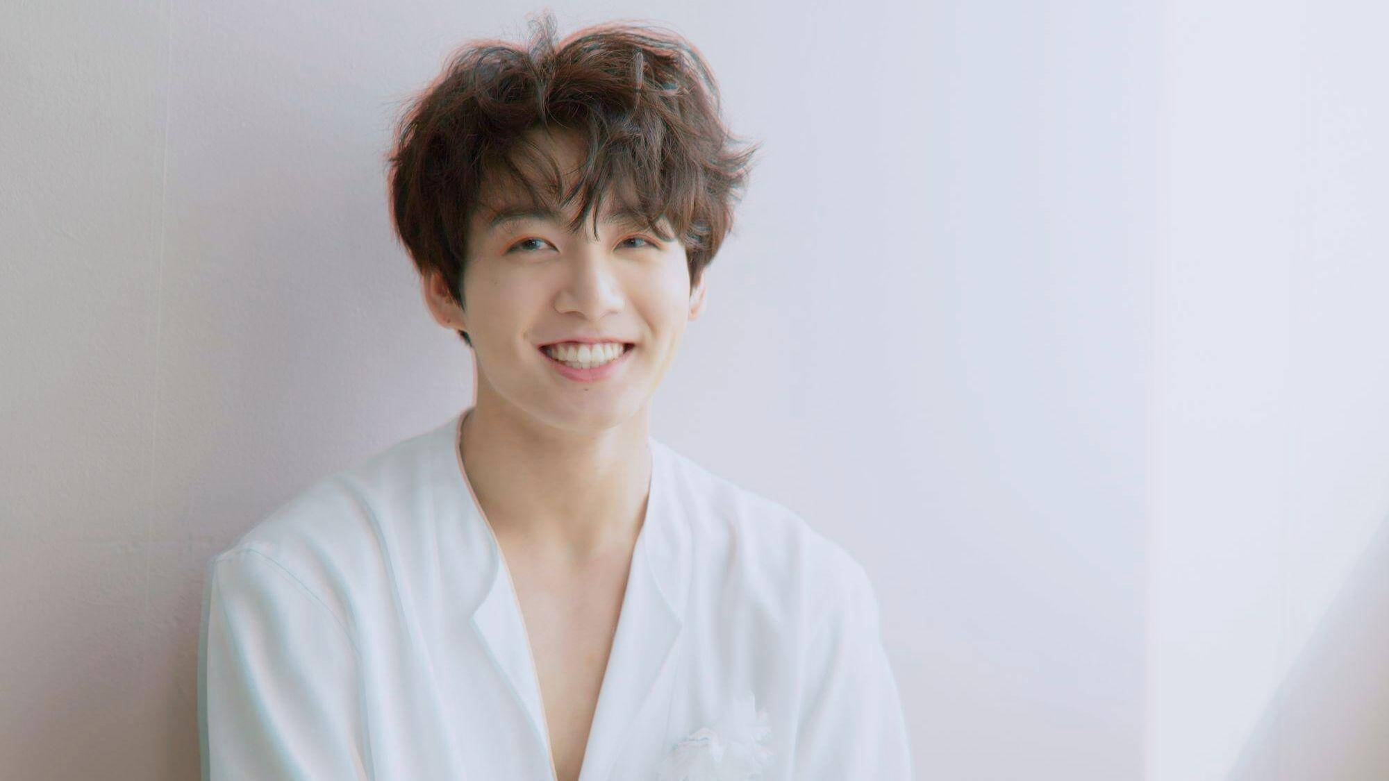 Jungkook 桌布 壁紙 — 一位年輕男子，棕色亂髮，穿著白襯衫，靠著柔軟的白牆，溫暖地微笑。