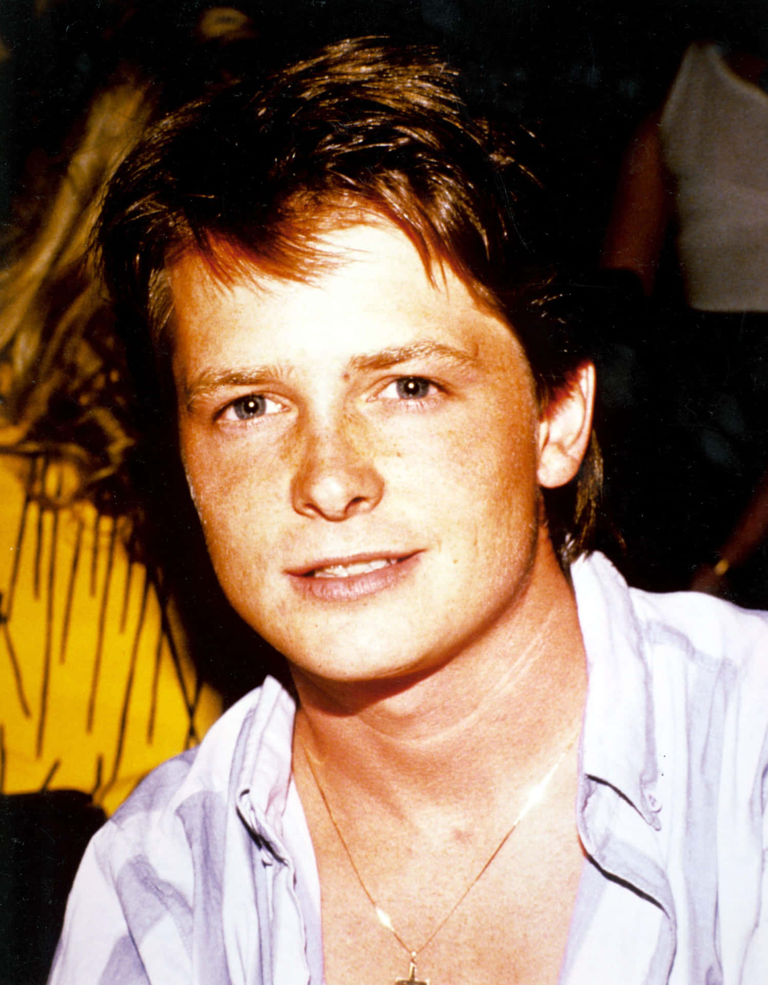 michael j fox 桌布 壁紙 — 一位年輕男子，棕色亂髮，臉上帶有雀斑，在昏暗的房間裡，黃色圖案背景前，溫柔地微笑。