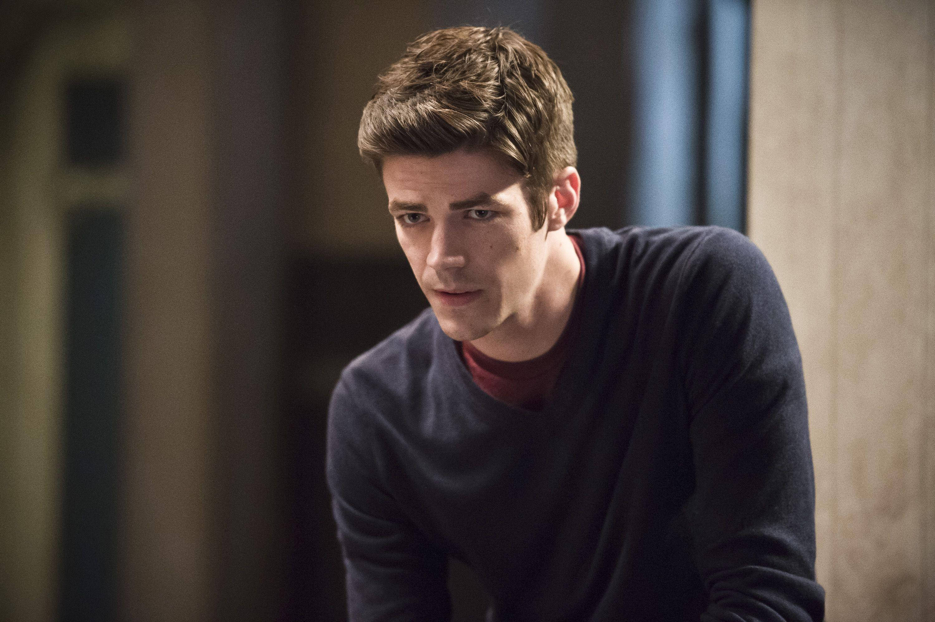 Grant Gustin 桌布 壁紙 — 一位年輕男子，棕色亂髮，身穿深色毛衣內搭紅色襯衫，在柔和光線的室內環境中專注地向前凝視。
