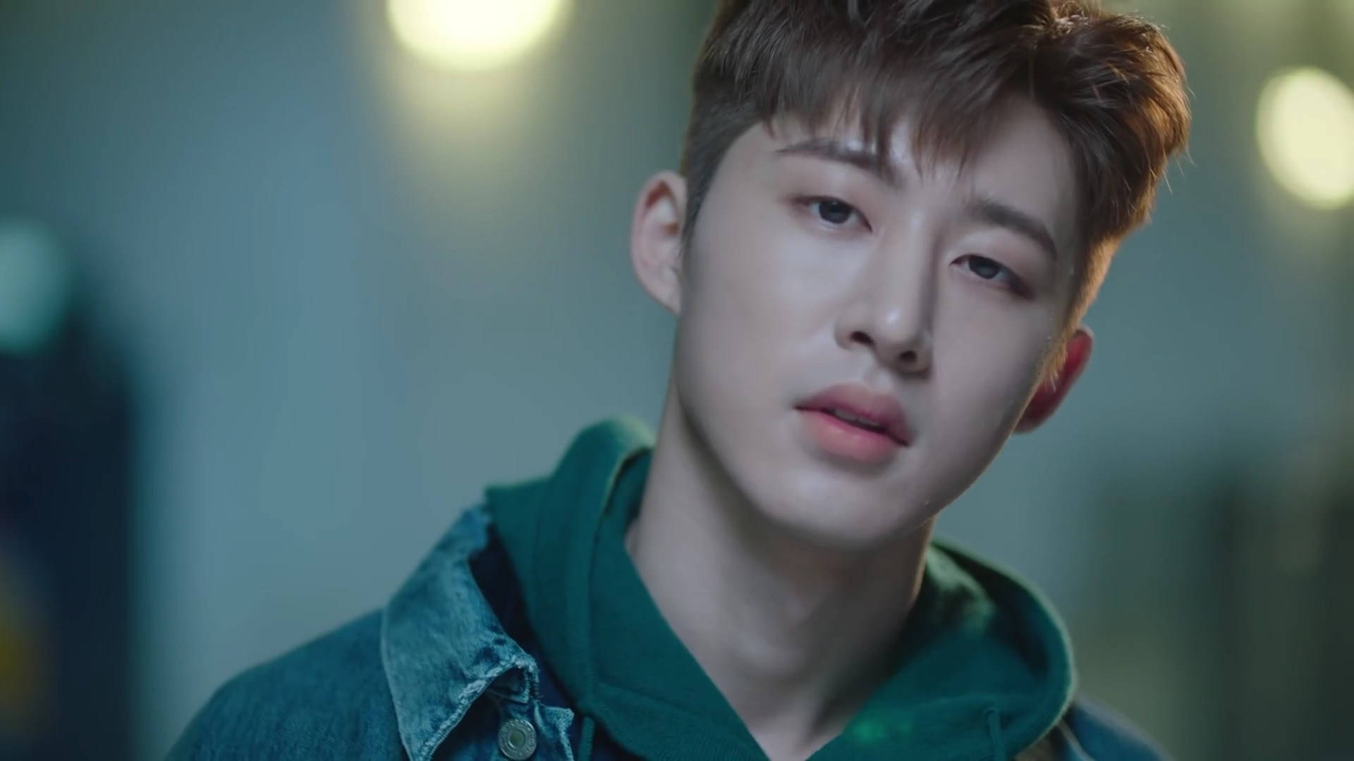 Ikon 桌布 壁紙 — 一位年輕男子，棕色亂髮，身穿綠色連帽衫外搭牛仔外套，在柔和模糊的背景和溫暖的散景燈光下若有所思地凝視。