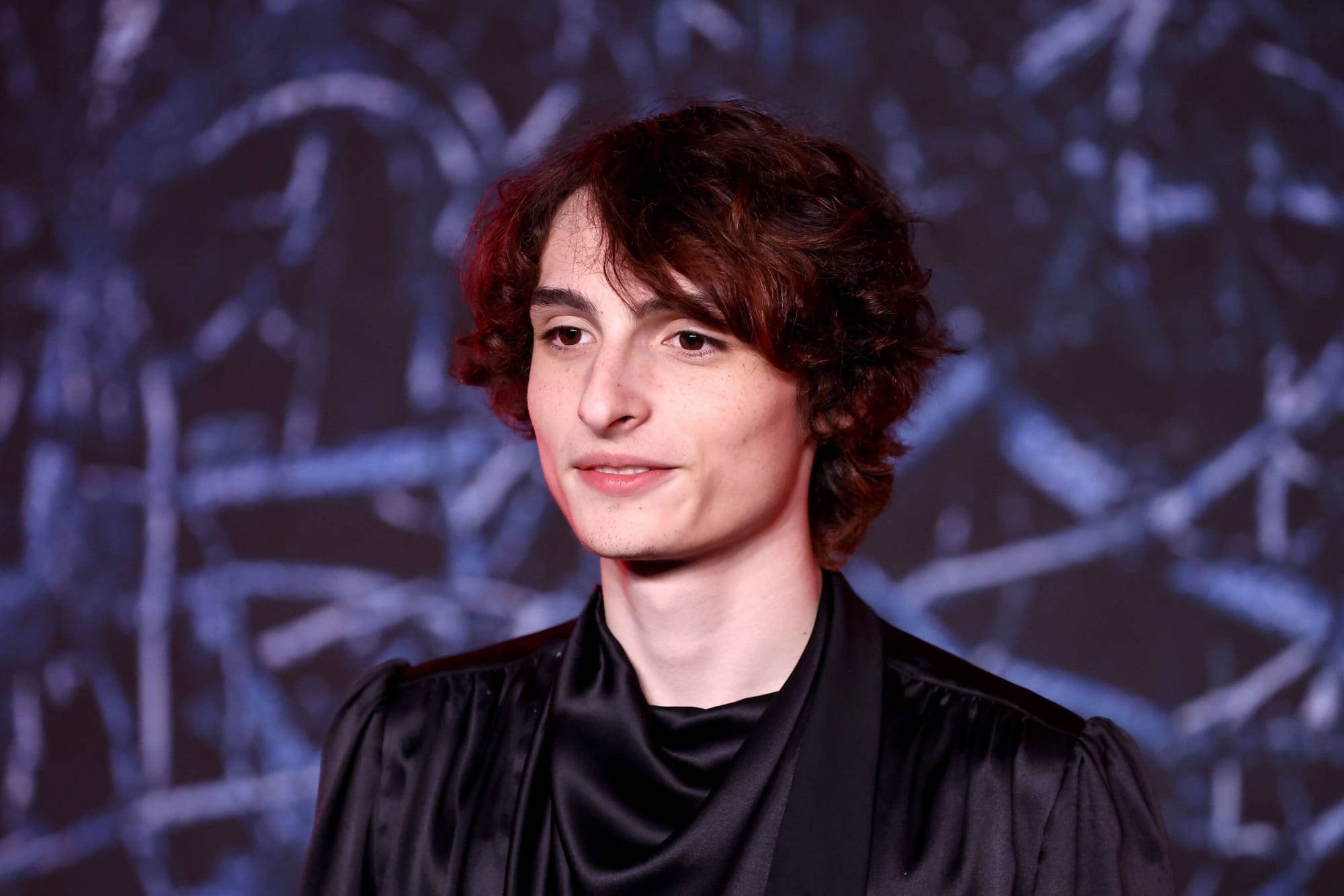 Celebrity 桌布 壁紙 — 一位年輕男子，棕色捲髮，身穿黑色緞面長袍，背景為藍色抽象圖案。