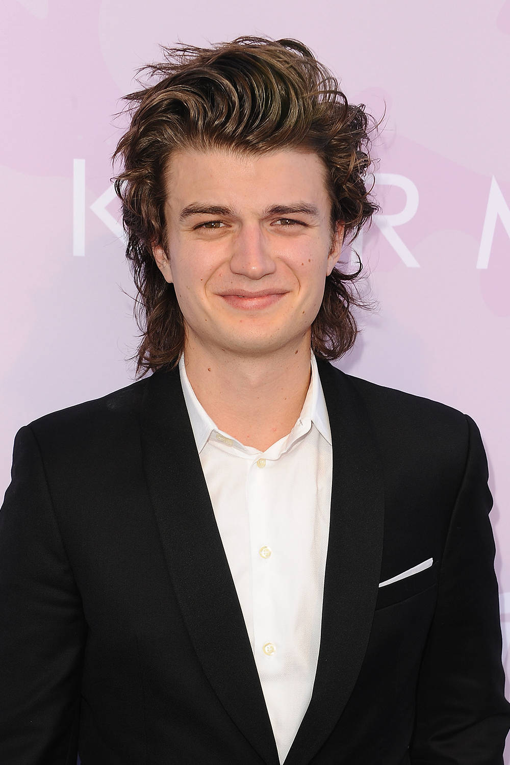 Joe Keery 桌布 壁紙 — 一位年輕男子，棕色頭髮略顯凌亂，身穿黑色西裝，在柔和的粉紅色背景下溫暖微笑。