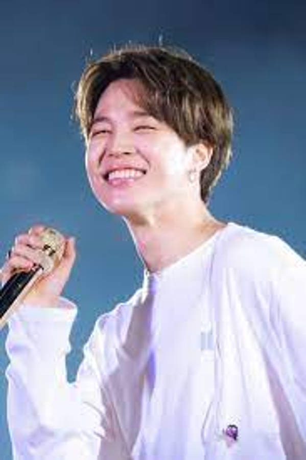BTS 智旻 桌布 壁紙 — 一位年輕男子，棕色頭髮略顯凌亂，在深藍色天空背景下，手持麥克風，笑容燦爛。
