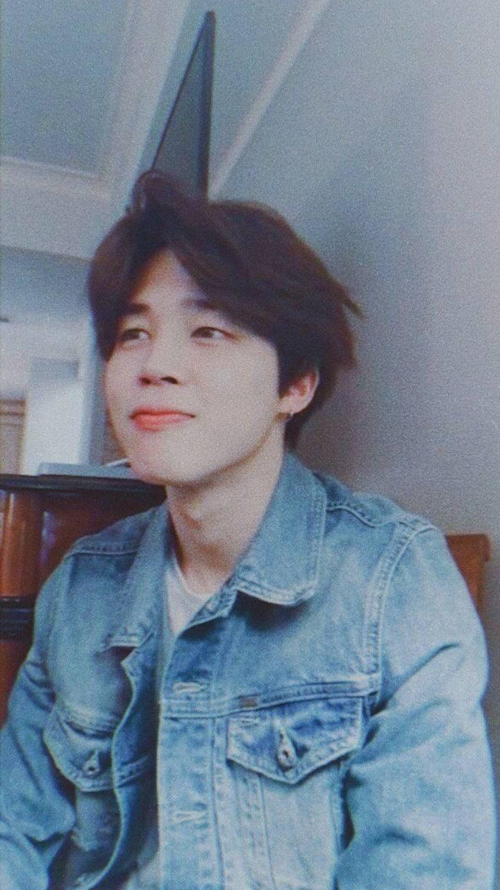 Jimin Aesthetic 桌布 壁紙 — 一位年輕男子，棕色頭髮略顯凌亂，在柔和的燈光下室內穿著牛仔外套，露出溫柔的微笑。
