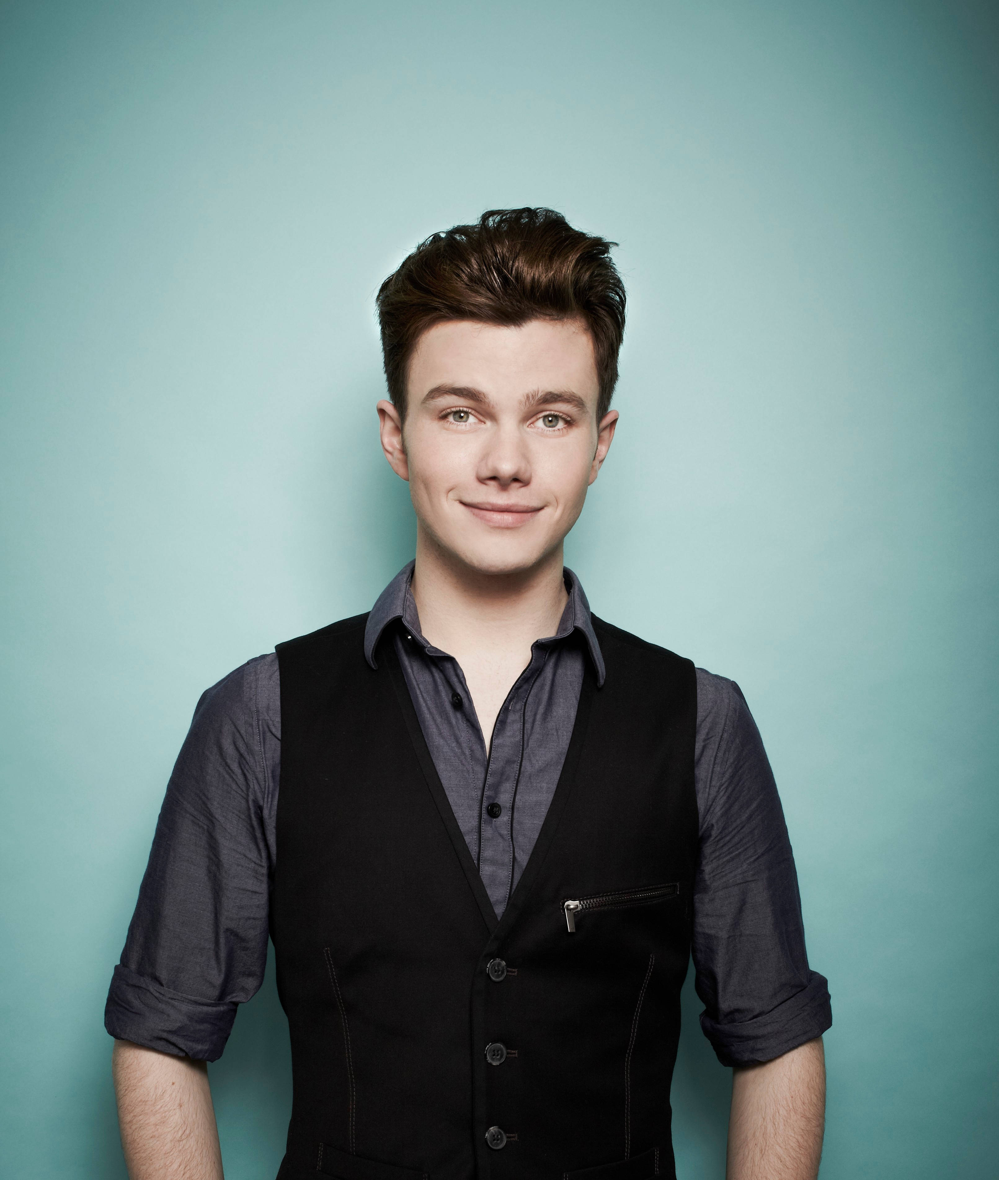 Chris Colfer 桌布 壁紙 — 一位年輕男子，棕色頭髮經過造型，在純色青綠色工作室背景前溫柔微笑，身穿灰色襯衫外搭黑色背心。