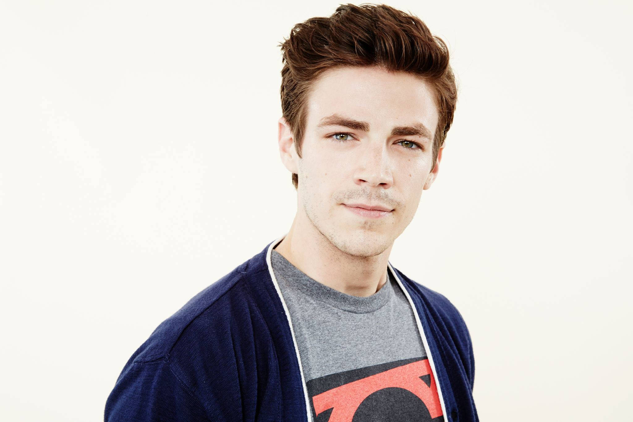 Grant Gustin 桌布 壁紙 — 一位年輕男子，棕色頭髮造型，帶著含蓄的微笑，身穿海軍藍開襟毛衣，內搭灰色T恤，上面有紅黑圖案，背景是純白色。