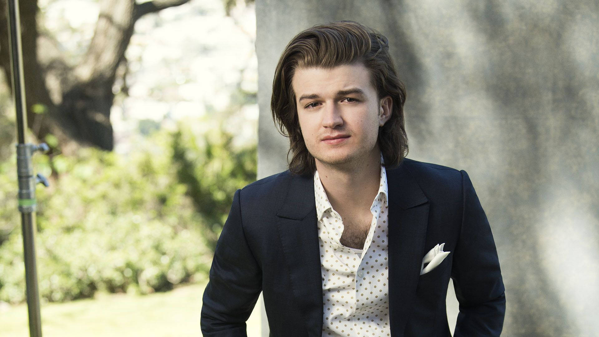Joe Keery 桌布 壁紙 — 一位年輕男子，棕色頭髮造型時尚，身穿深色西裝外套，內搭白色圓點襯衫，背景是陽光普照的花園。