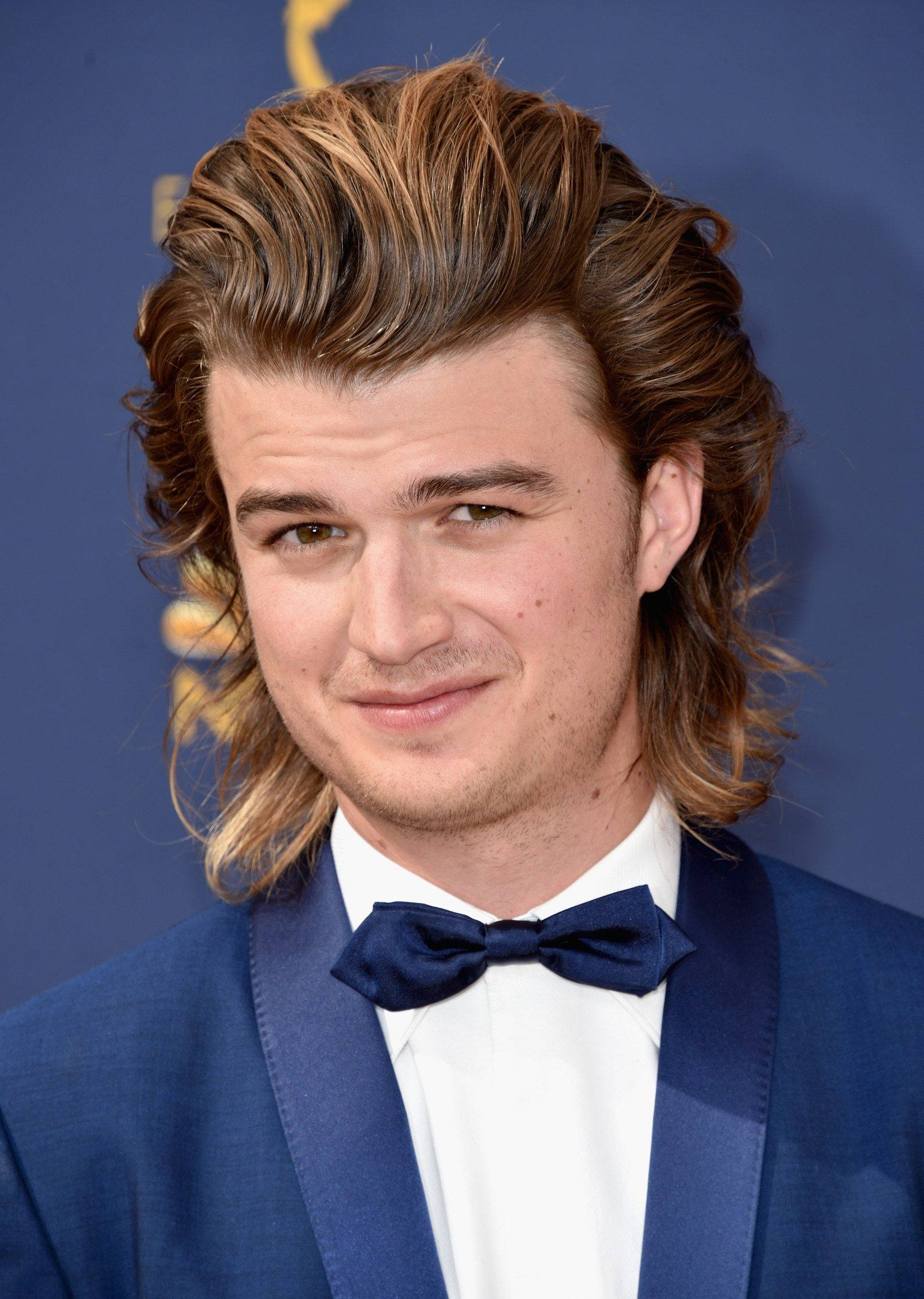Joe Keery 桌布 壁紙 — 一位年輕男子，棕色頭髮造型時尚，身穿藍色燕尾服和領結，背景為藍色，帶有金色艾美獎標誌。