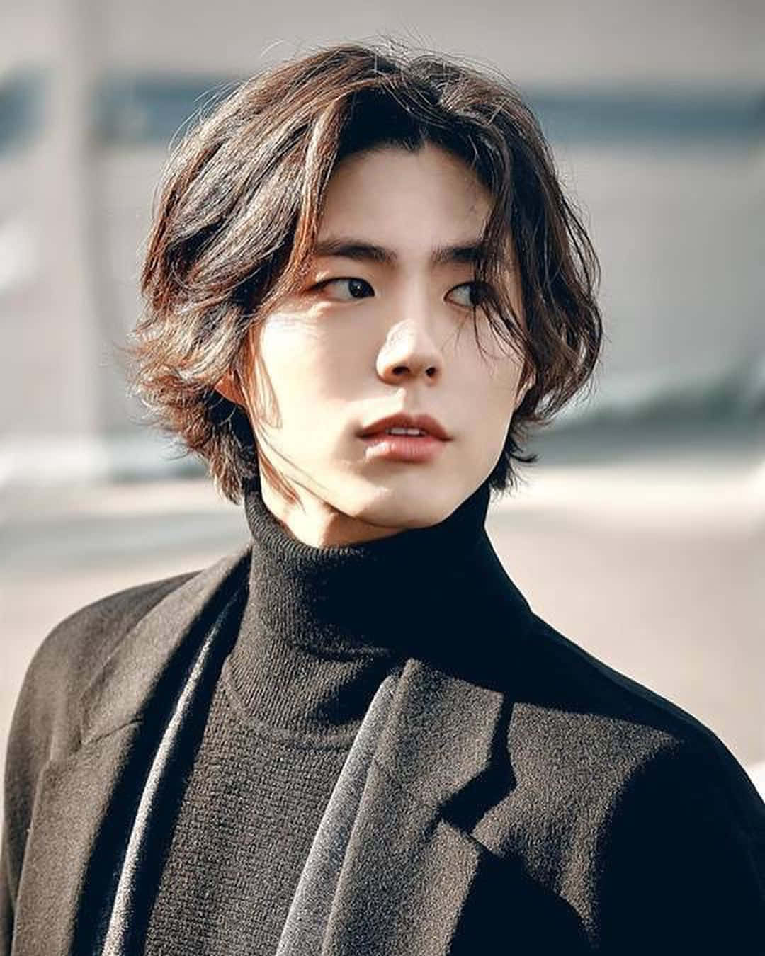 Hairstyle 桌布 壁紙 — 一位年輕男子，波浪形棕色頭髮，穿著黑色高領毛衣和大衣，在柔和的日光下，背景是模糊的城市景觀。