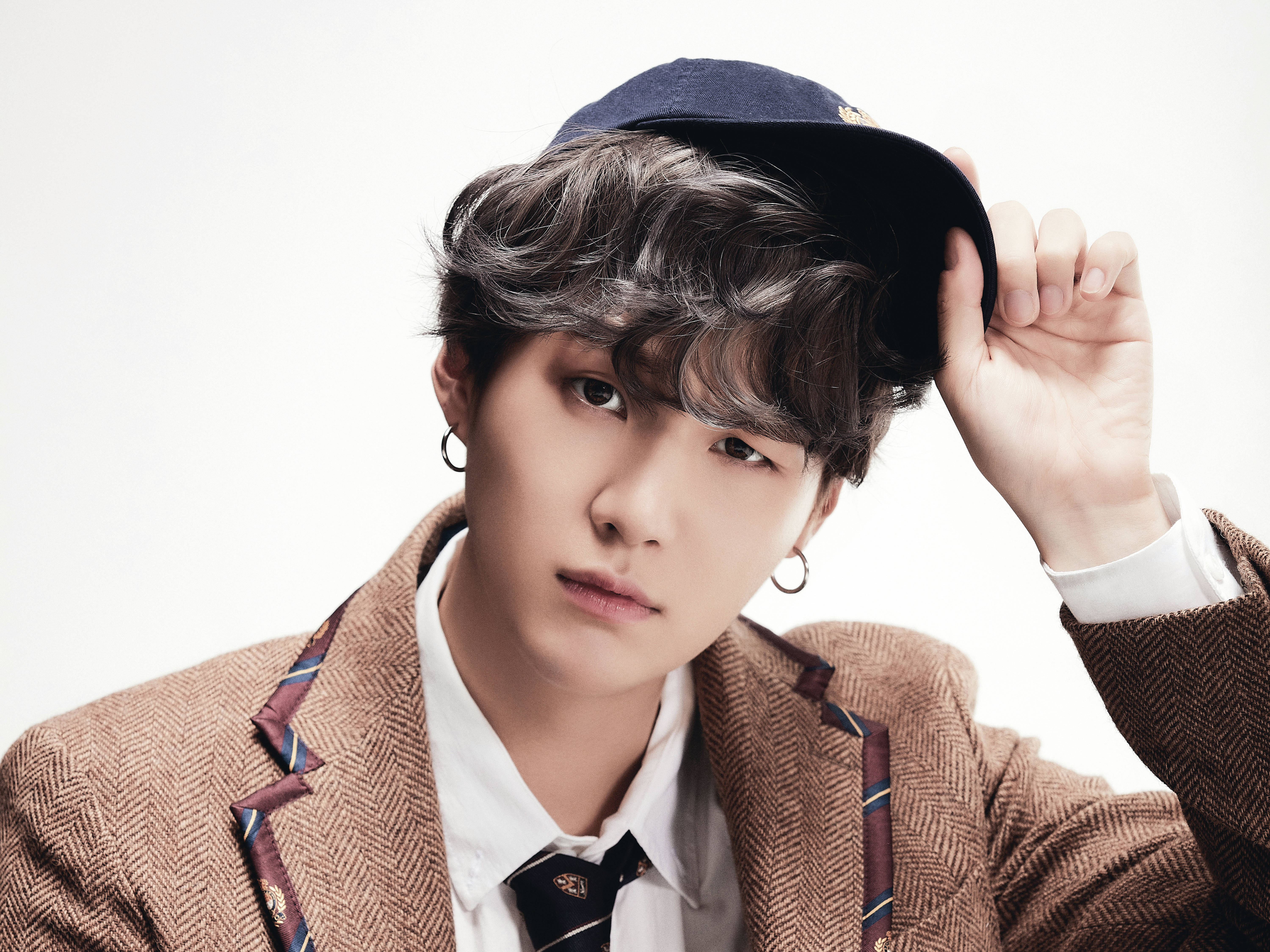 Suga Bts 桌布 壁紙 — 一位年輕男子，波浪頭髮戴著耳環，身穿棕色人字紋西裝外套，內搭白襯衫和圖案領帶，正在調整海軍藍帽子，背景為純白色。