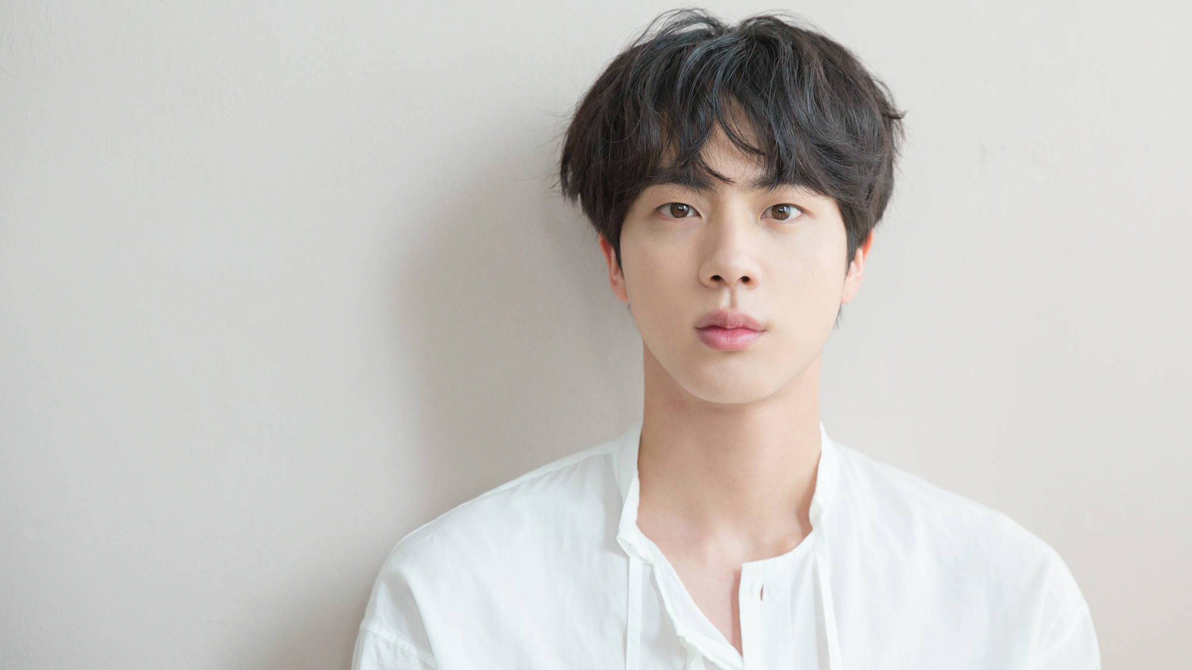 Jin 桌布 壁紙 — 一位年輕男子，深色凌亂的頭髮，表情平靜，穿著一件挺括的白襯衫，背景是純色的米白色牆壁。