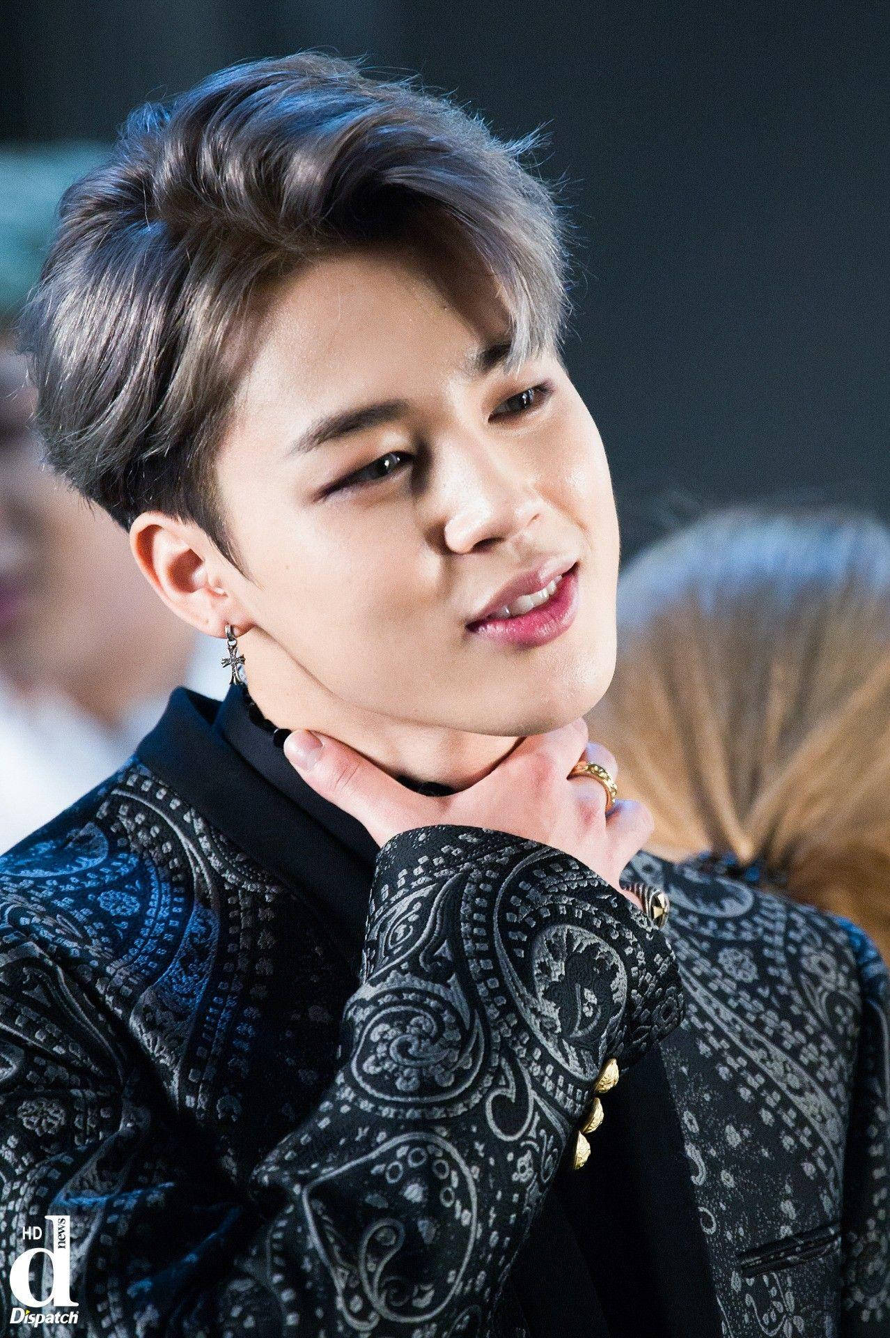 Jimin Bts Cute 桌布 壁紙 — 一位年輕男子，留著時尚髮型並戴著耳環，穿著帶有圖案的黑色外套，輕輕觸摸自己的頸部。