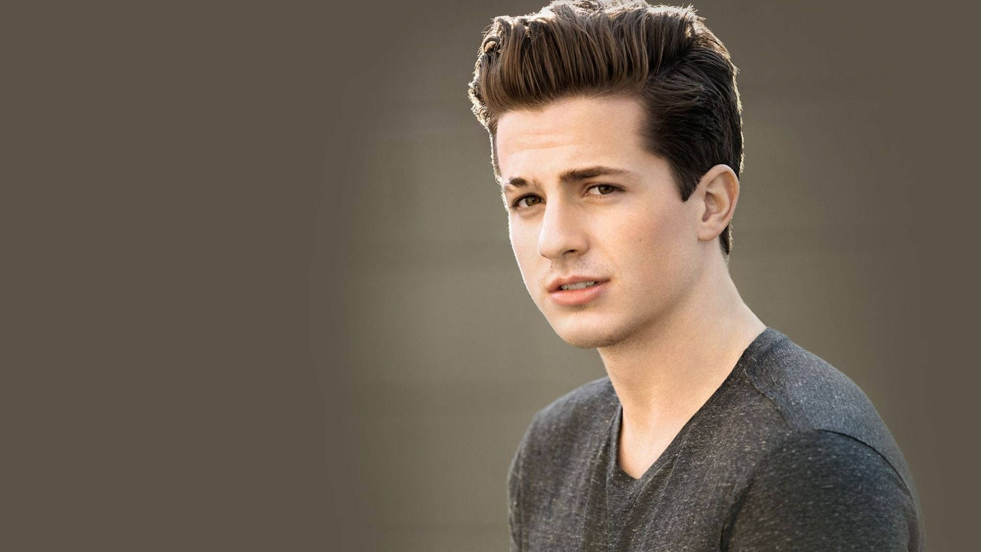 Charlie Puth 桌布 壁紙 — 一位年輕男子，留著造型棕色頭髮，身穿灰色V領襯衫，在柔和的灰色背景前若有所思地凝視。