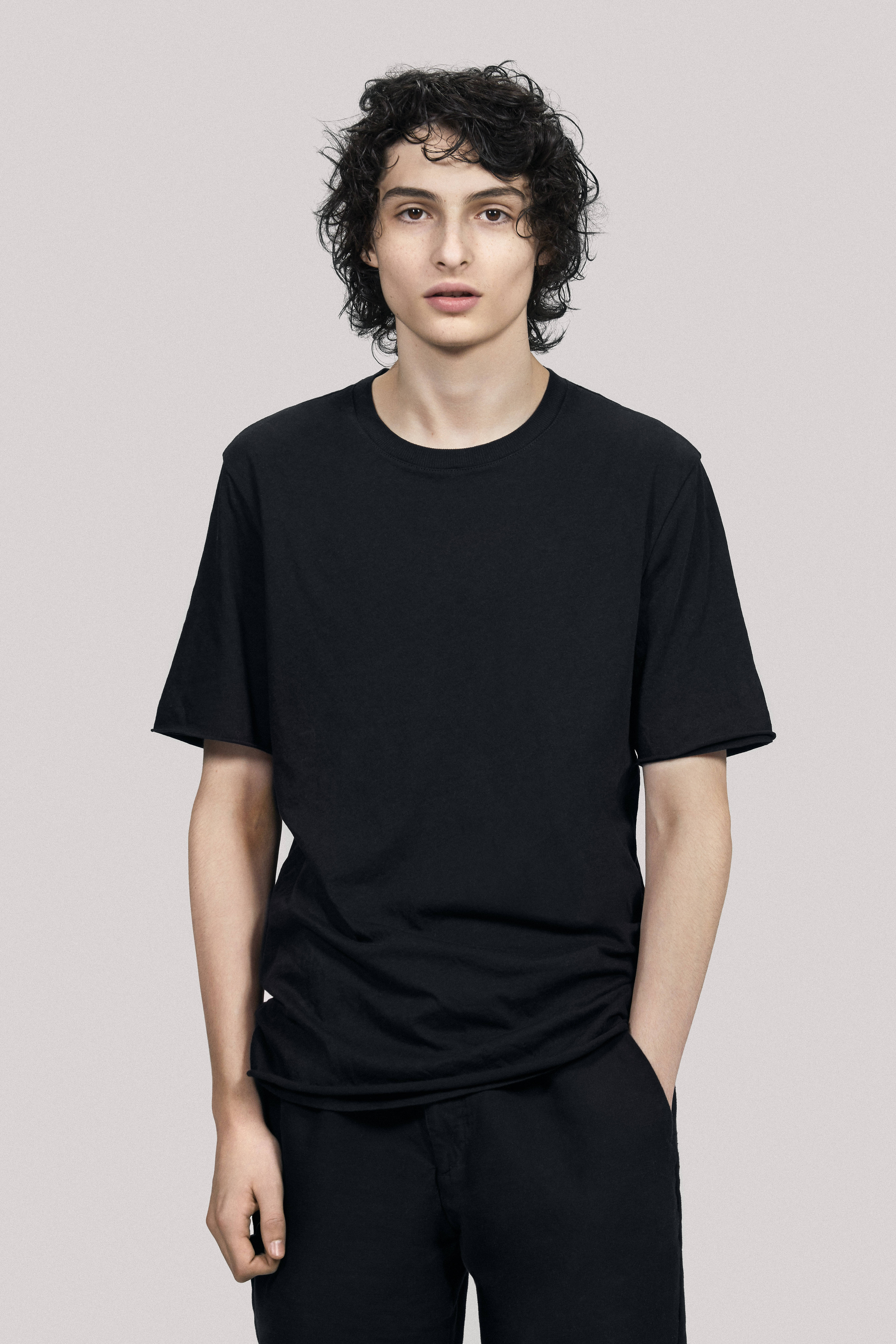 finn wolfhard 桌布 壁紙 — 一位年輕男子，留著黑色捲髮，身穿純黑色T恤和黑色長褲，背景為中性灰色。