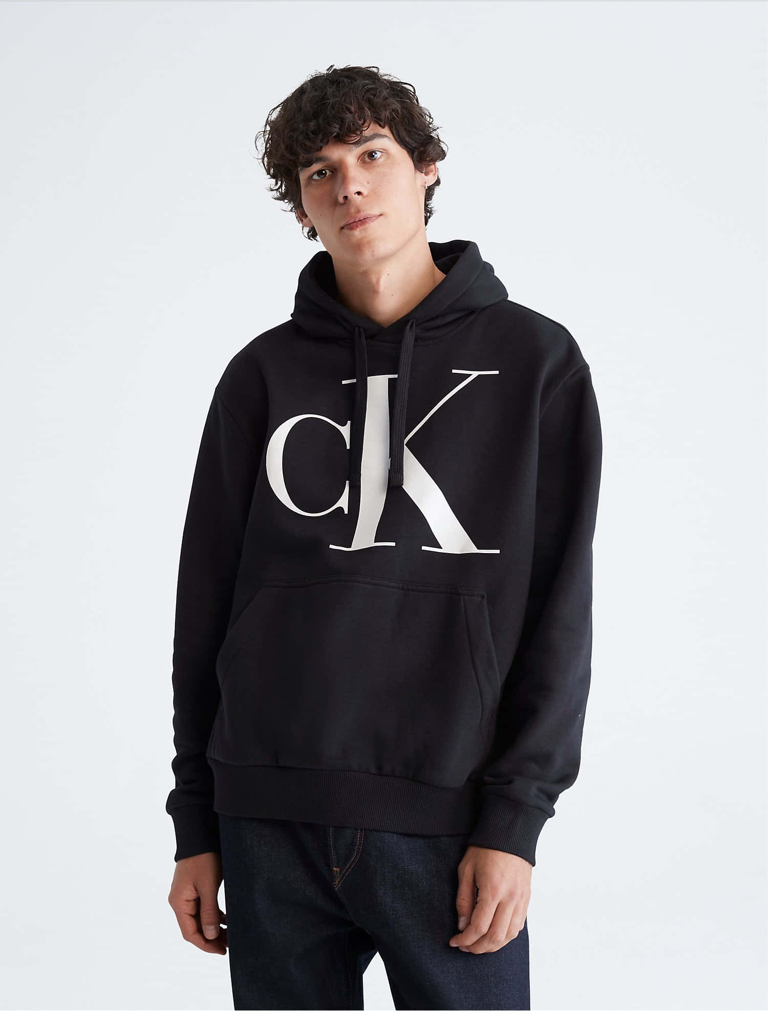 卡文克萊 桌布 壁紙 — 一位年輕男子，留著黑色捲髮，身穿黑色Calvin Klein連帽衫，胸前有大型白色標誌，背景為純白色。