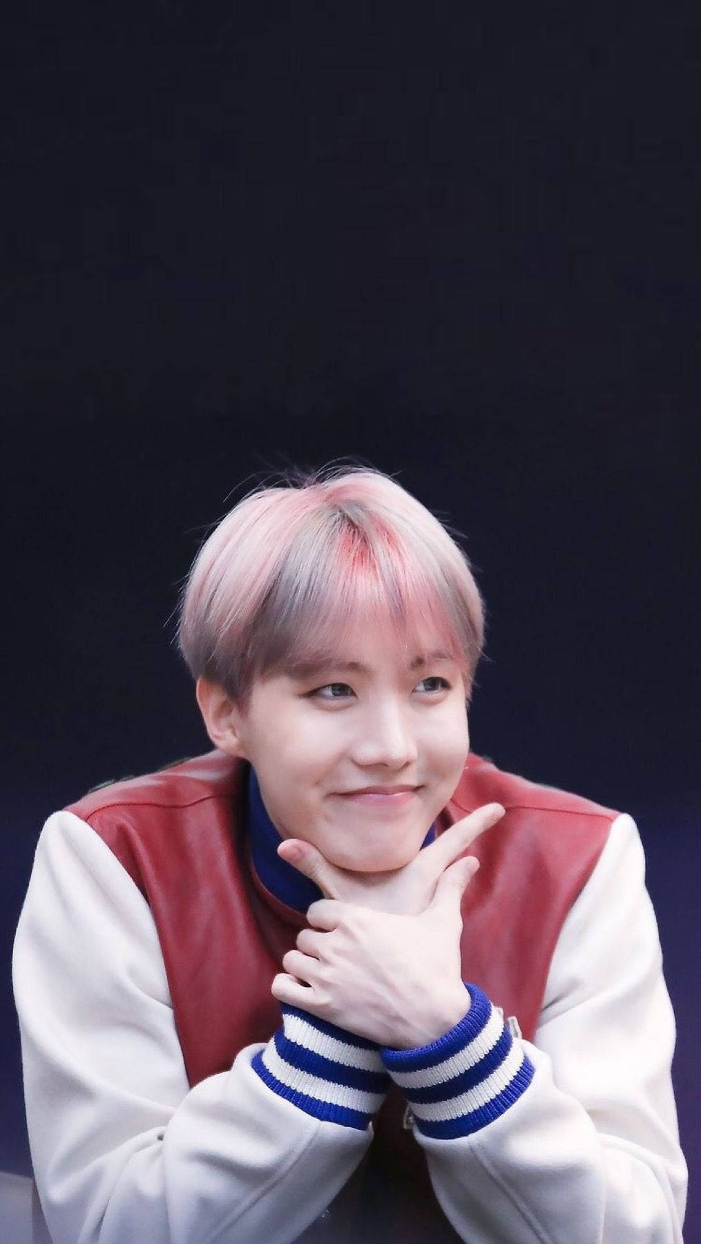 jhope 桌布 壁紙 — 一位年輕男子，粉紅色和灰色頭髮，在深色背景上用手托著下巴，溫柔地微笑。