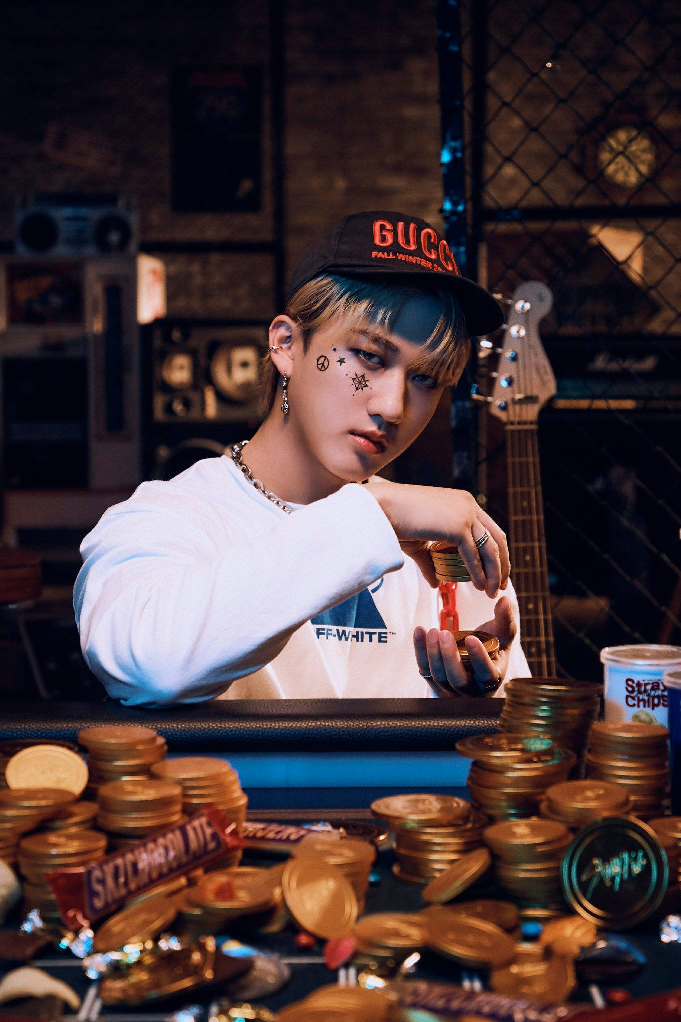 Stray Kids 桌布 壁紙 — 一位年輕男子，身上有星星刺青，戴著Gucci帽子，在一個光線昏暗、擺放著吉他和復古裝飾的房間裡，俯身在一張堆滿金幣和糖果的桌子上。