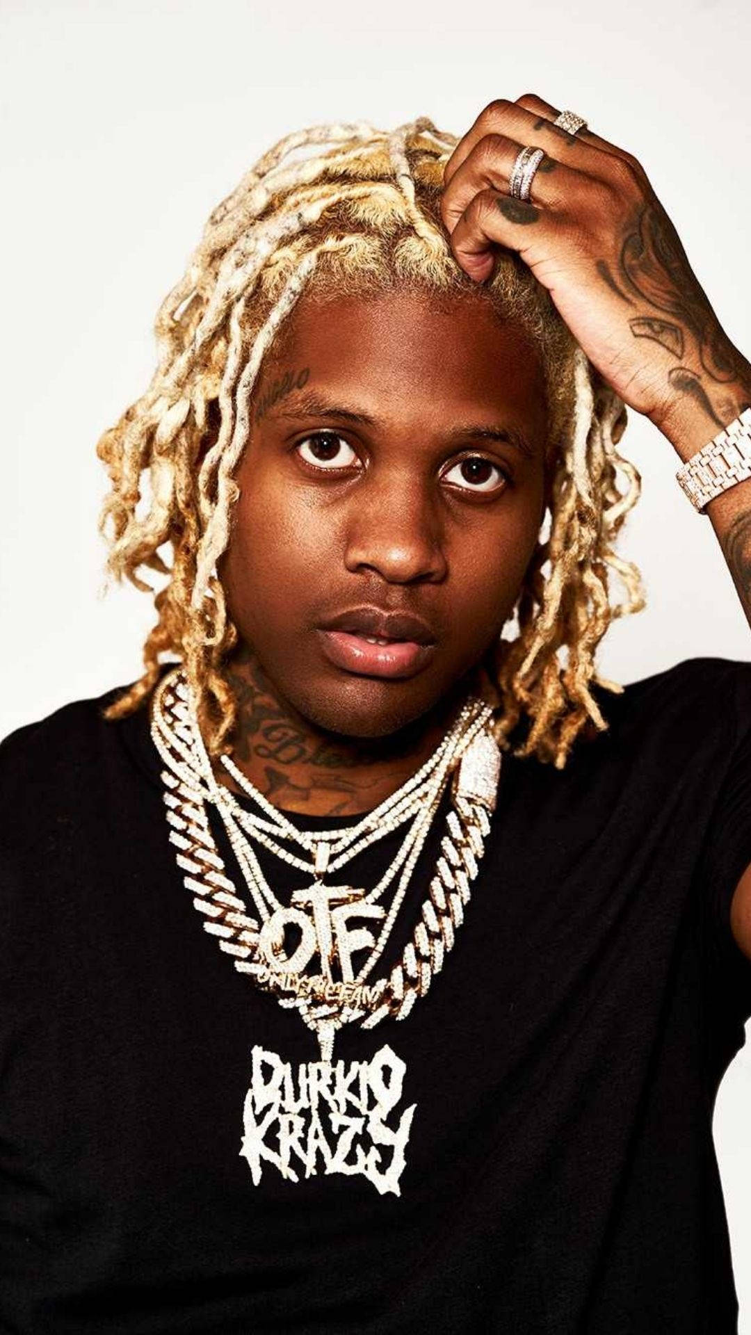 Lil Durk 桌布 壁紙 — 一位年輕男子，金色辮子頭和刺青，自信地擺姿勢，戴著多層鑽石鏈條和黑色圖案T恤，背景是純白色。