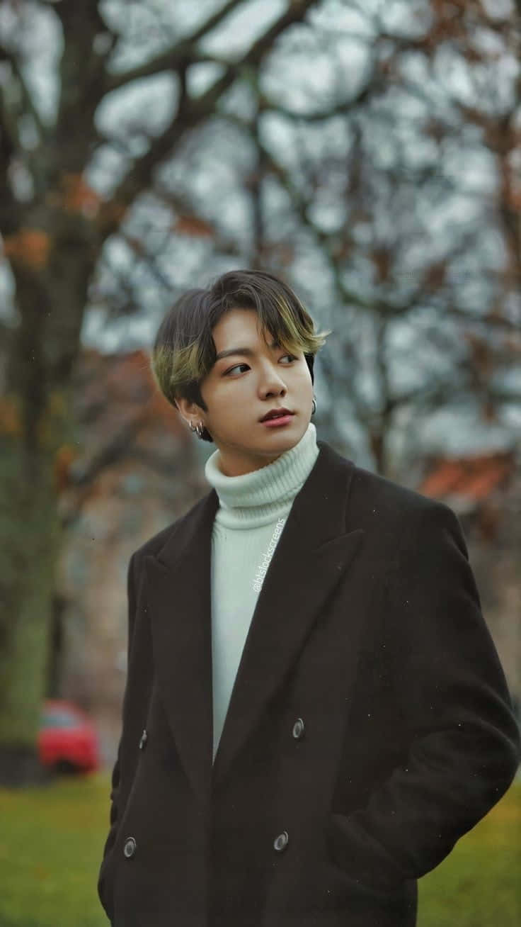 Jungkook 桌布 壁紙 — 一位年輕男子，頭髮造型時尚，戴著耳環，身穿黑色外套內搭白色高領毛衣，站在公園裡，背景是光禿的樹木和柔和的秋色。