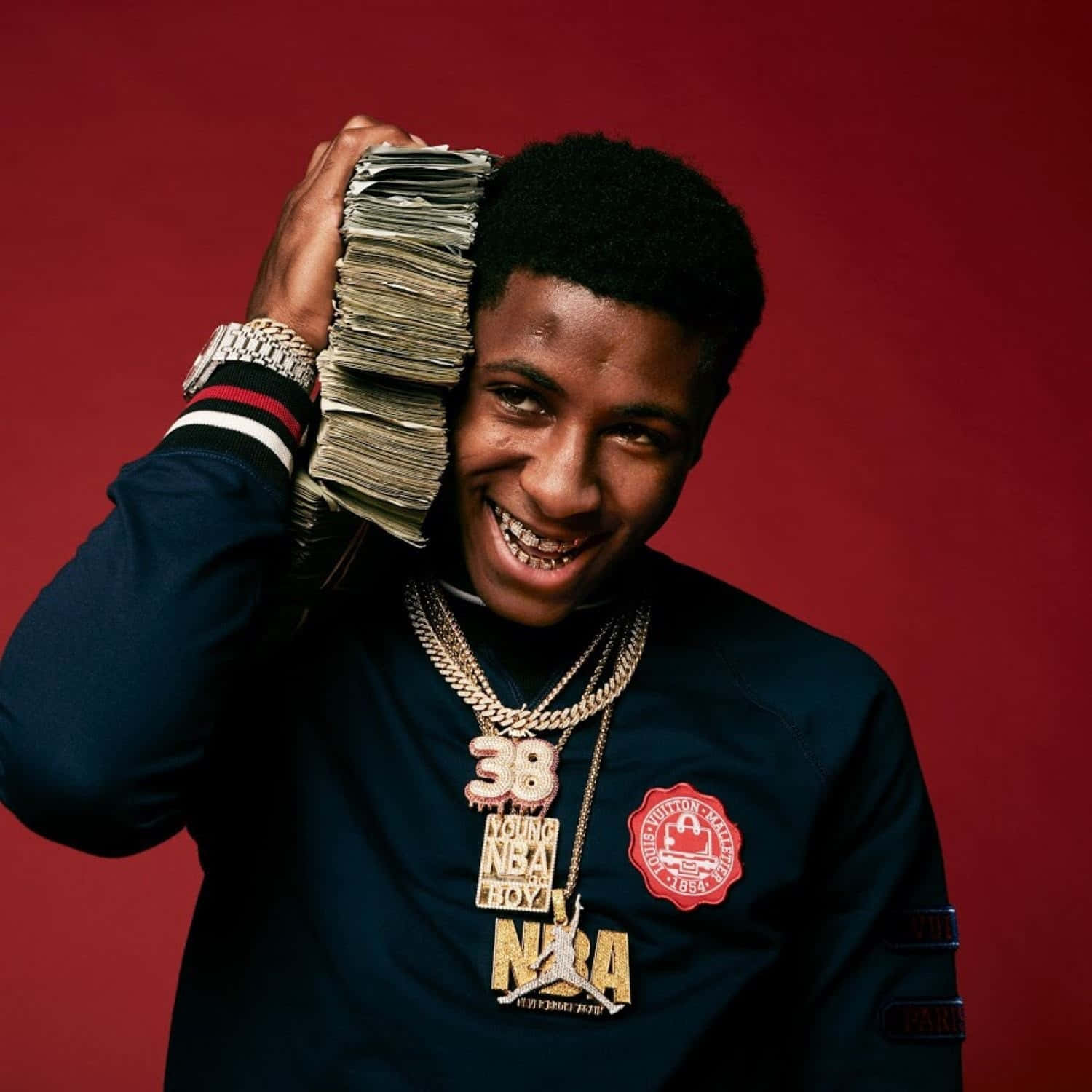 Youngboy 桌布 壁紙 — 一位年輕男子咧嘴大笑，將一疊厚厚的現金貼在頭上，身穿閃亮珠寶和品牌外套，背景為深紅色。
