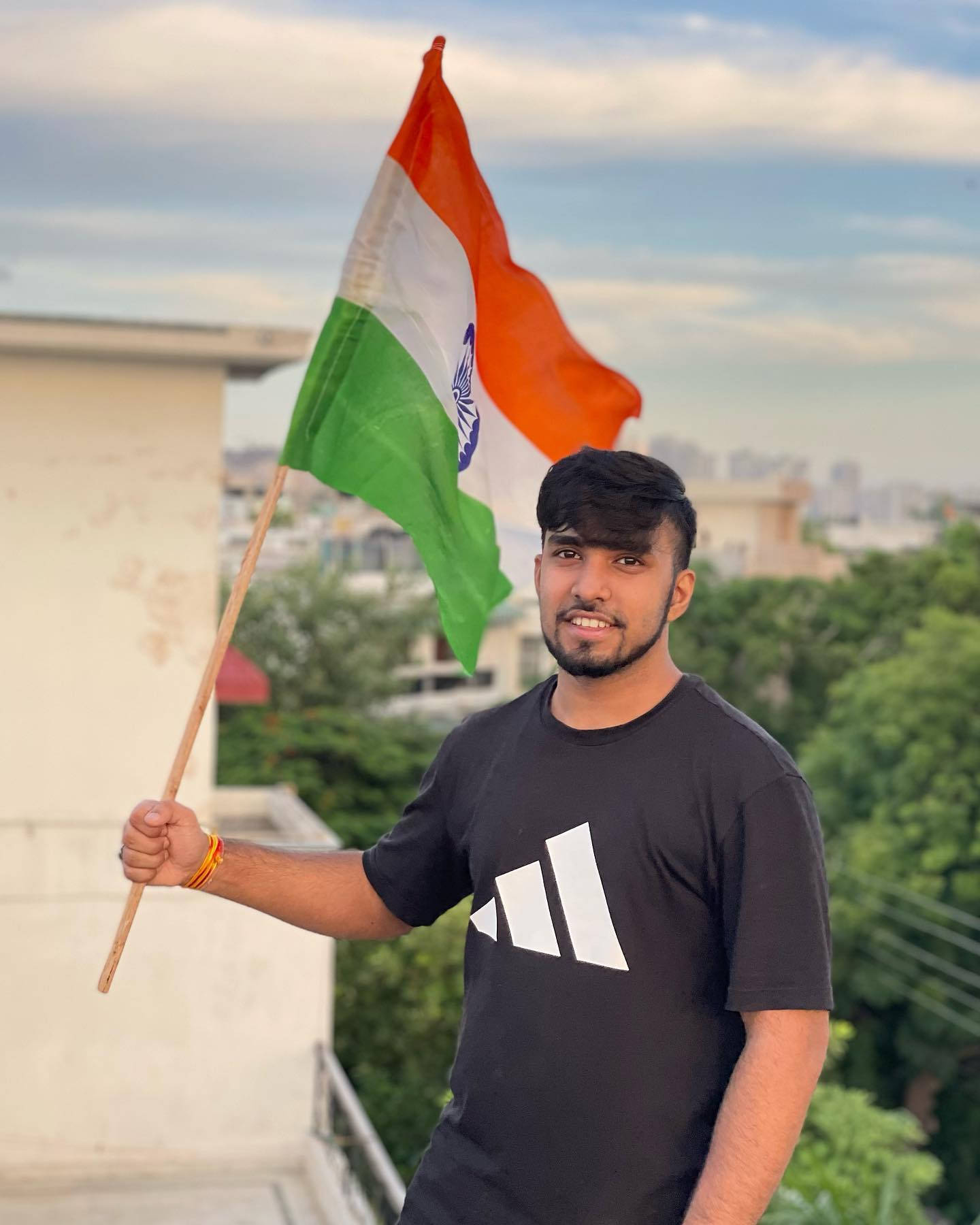 Indian 桌布 壁紙 — 一位年輕男子在屋頂上自豪地舉起印度國旗，背景是城市景觀和綠樹，天空部分多雲。