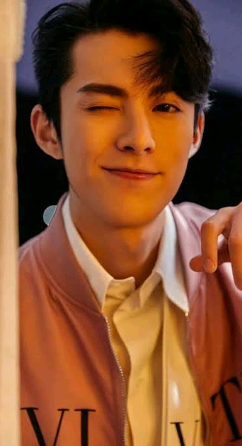 Dylan Wang 桌布 壁紙 — 一位年輕男子在溫暖的金色燈光下，穿著粉紅色飛行員夾克，頑皮地眨眼。