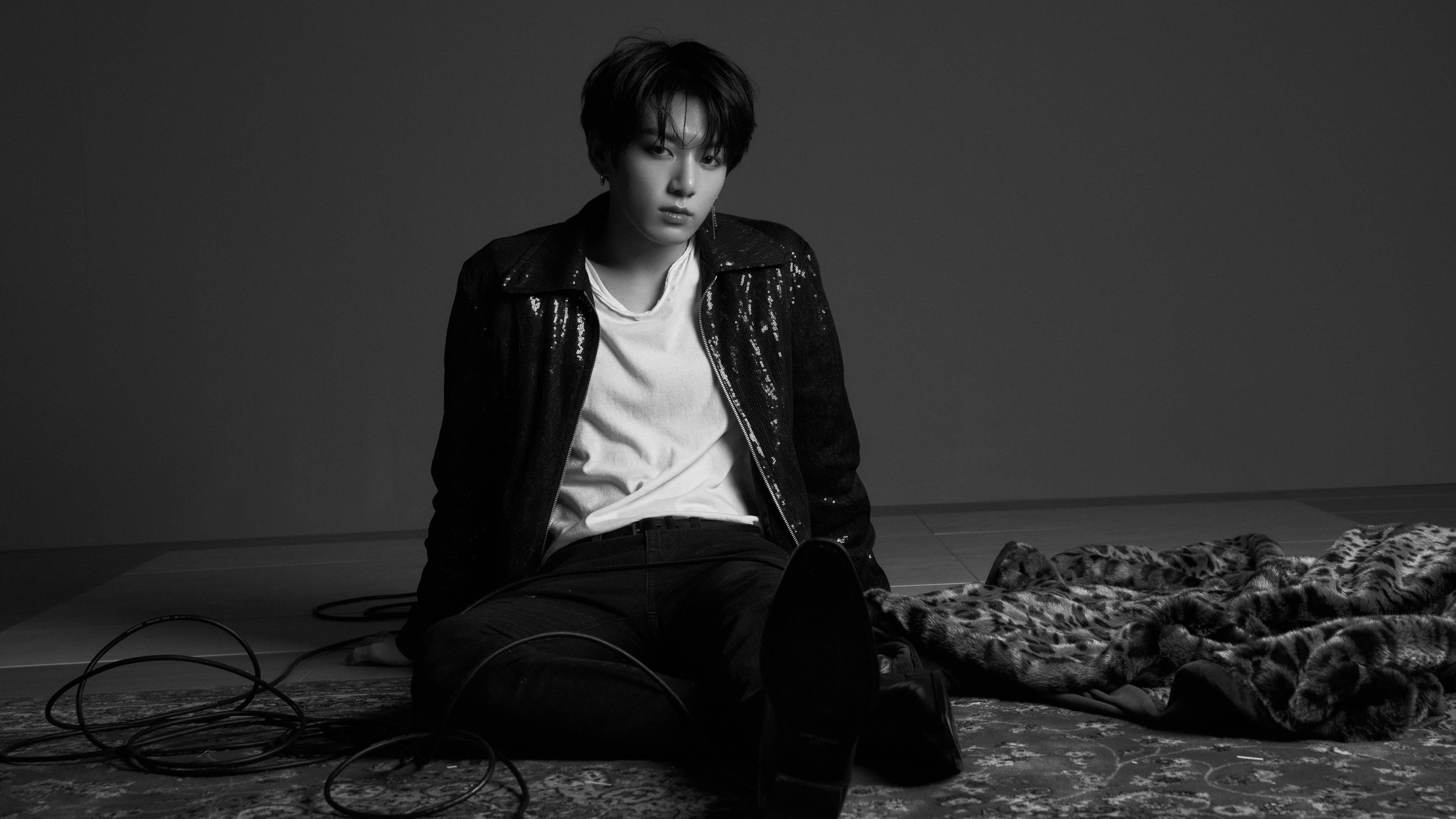Bts Black 桌布 壁紙 — 一位年輕男子坐在簡約工作室的圖案地毯上，身穿白色T恤外搭閃亮黑色外套，周圍環繞著纏繞的電線和質感毛毯。