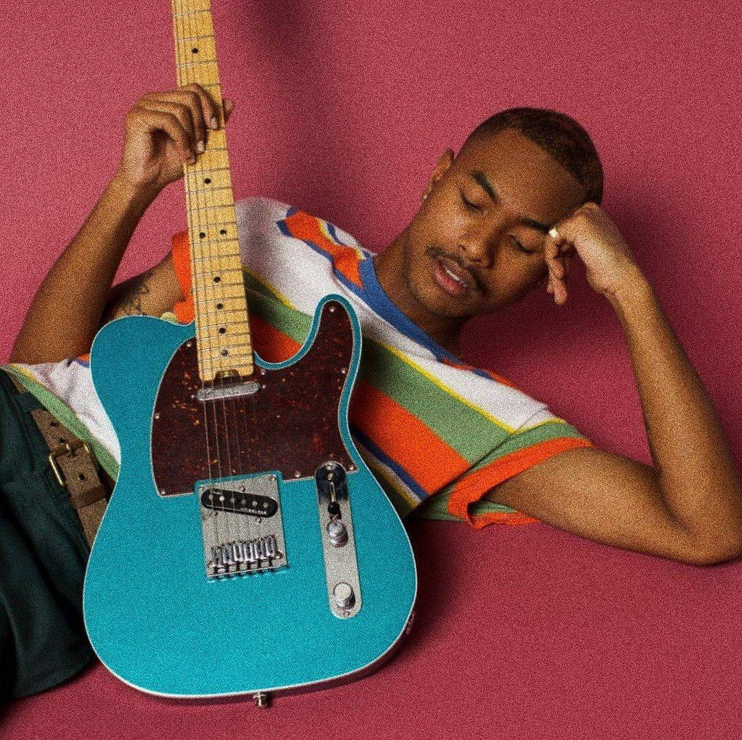 Steve Lacy 桌布 壁紙 — 一位年輕男子將頭靠在鮮豔的粉紅色表面上，同時手持一把藍綠色電吉他。