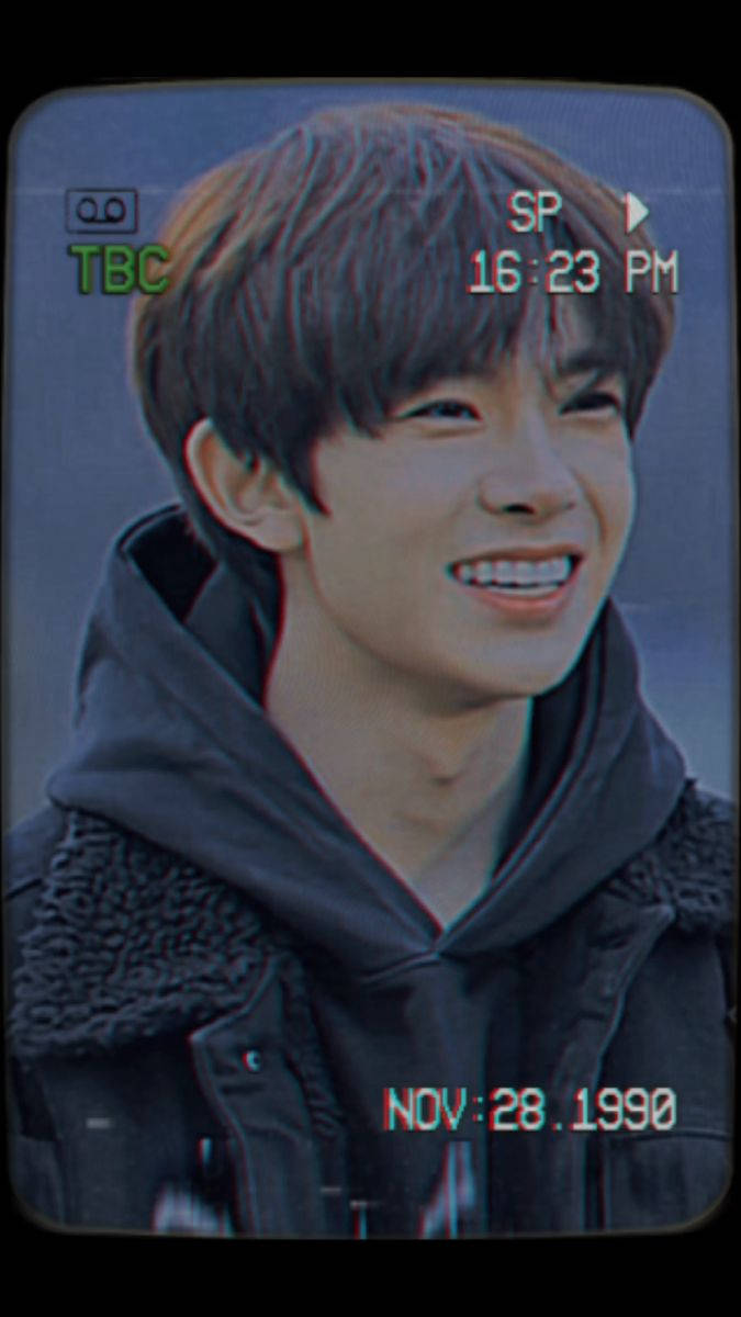 Kpop Aesthetic 桌布 壁紙 — 一位年輕男子帶著燦爛笑容，在模糊背景前穿著深色連帽衫，外搭人造毛邊夾克。