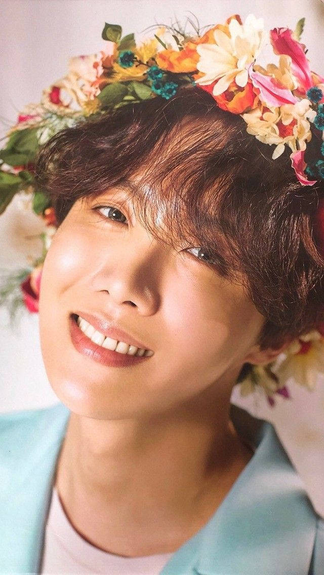 j-hope 桌布 壁紙 — 一位年輕男子帶著燦爛笑容，戴著色彩繽紛的花冠，背景是柔和的粉彩色。